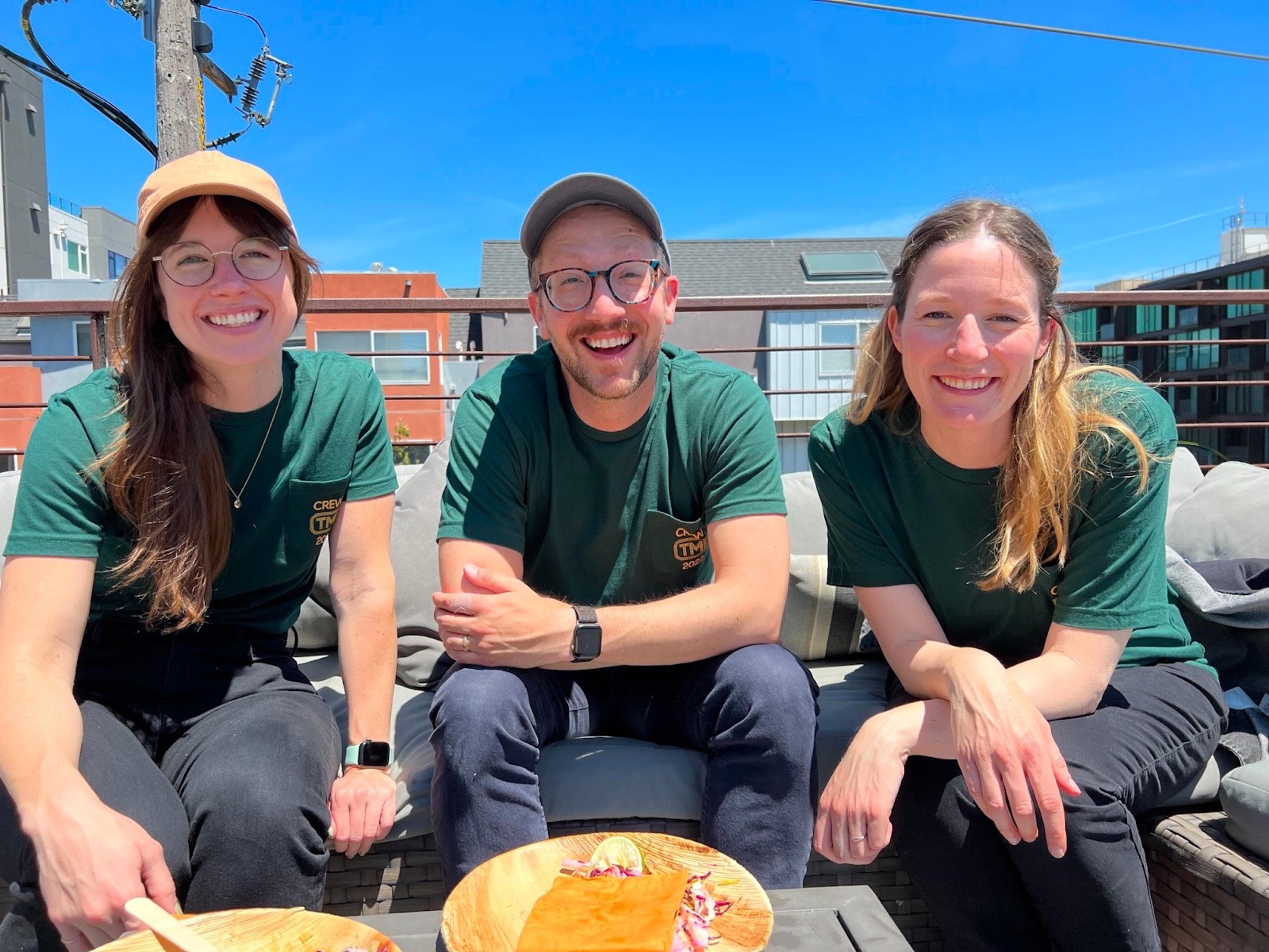 Three people sitting and smiling happily at the camera. They are all wearing dark green t-shirts and black jeans. The woman on the left wears glasses and a tan cap. The man in the middle sports a cap and glasses as well as short facial hair. The woman on the right has long blonde hair tucked behind her ears.