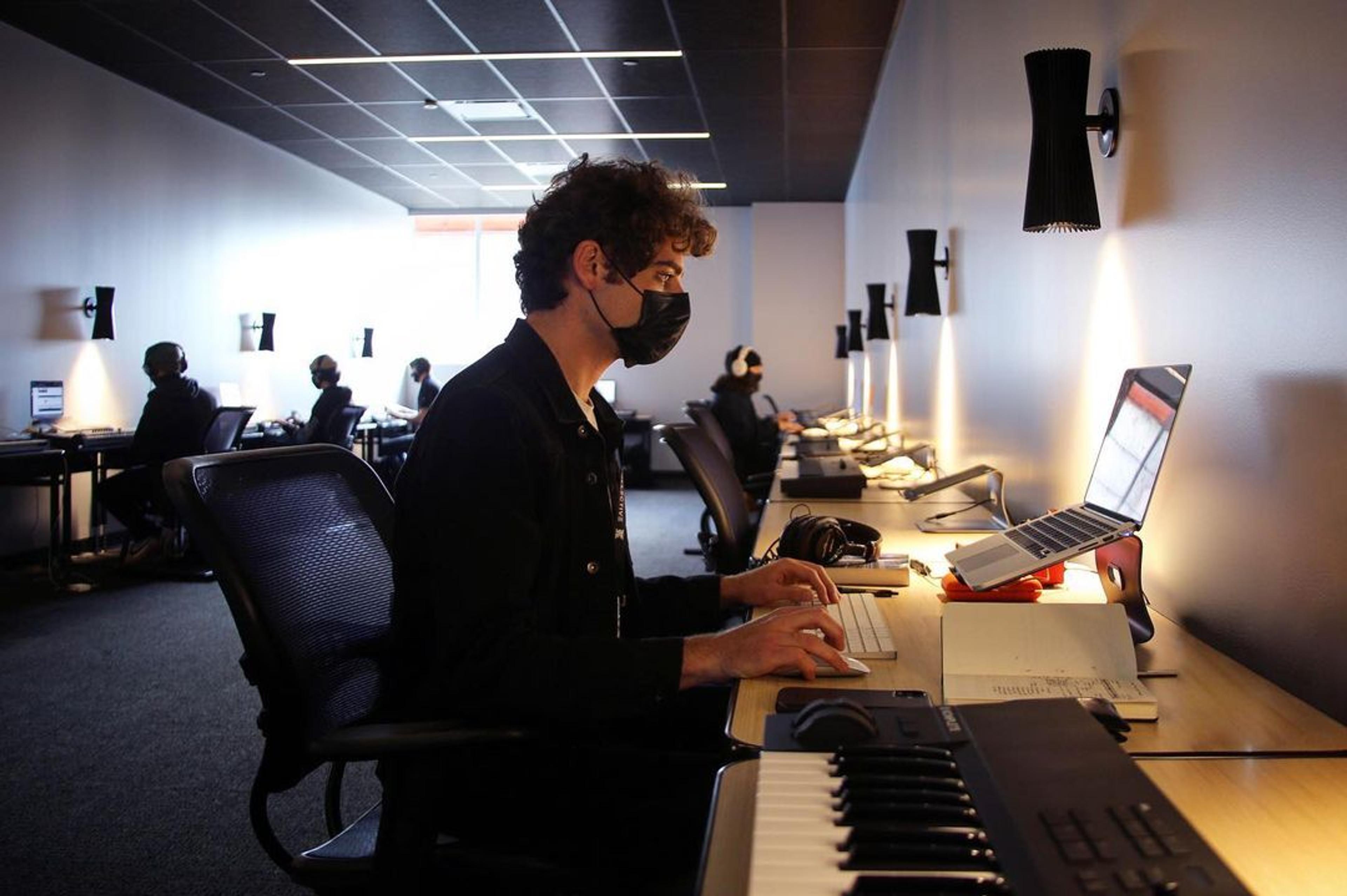 A photo of a man wearing a mask and sitting at a desk working on a laptop. Next to the laptop is a digital MIDI keyboard,