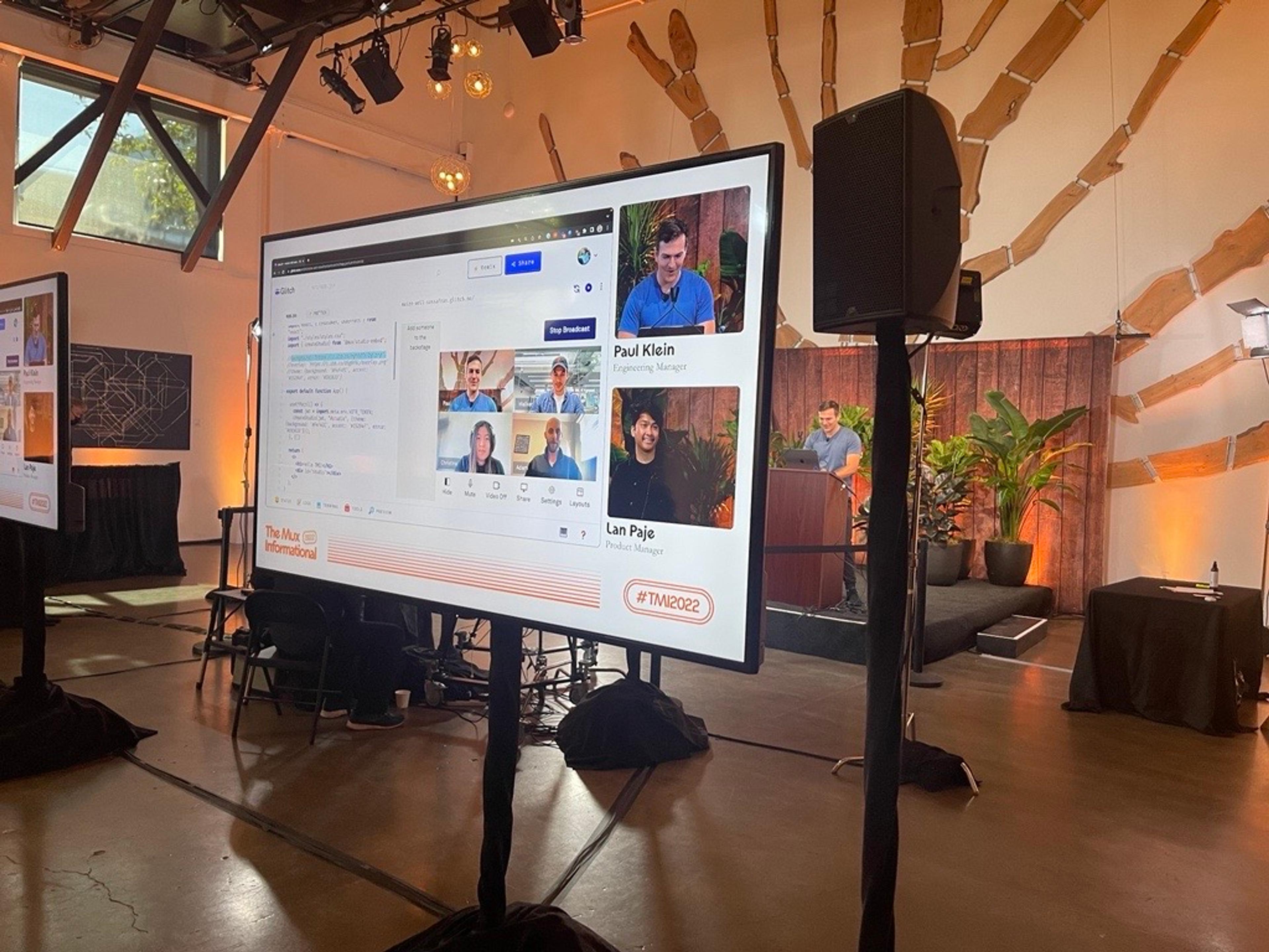 A backstage look at a man named Paul Klein standing behind a podium on stage wearing a blue shirt along with another man named Lan Paje, wearing a black shirt. They are both visible, along with their presentation slides, on a monitor in the foreground.