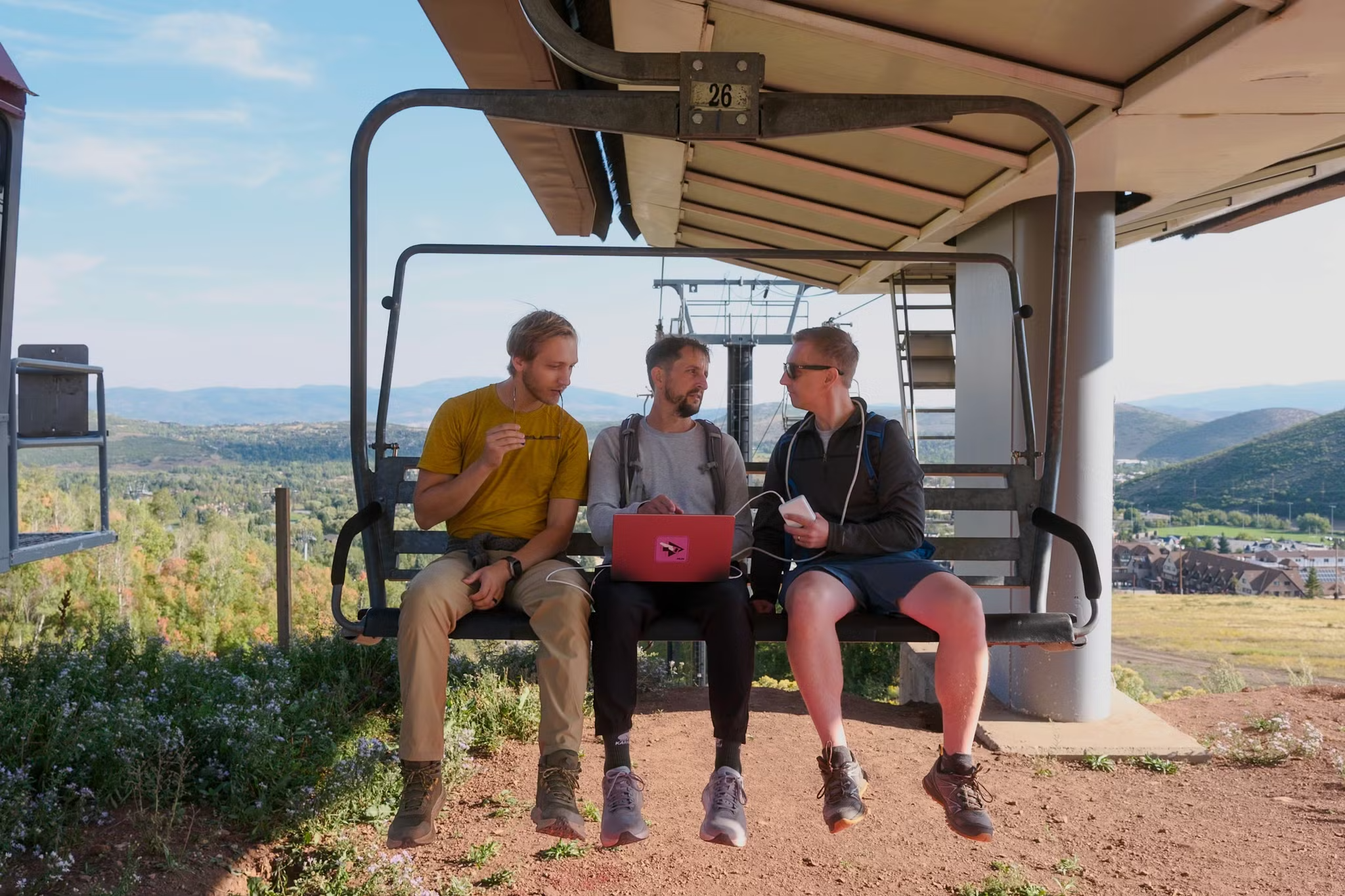 Rob, Dar, and Dave conversing while they sit on a chair lift. Dave has a laptop in his lap.