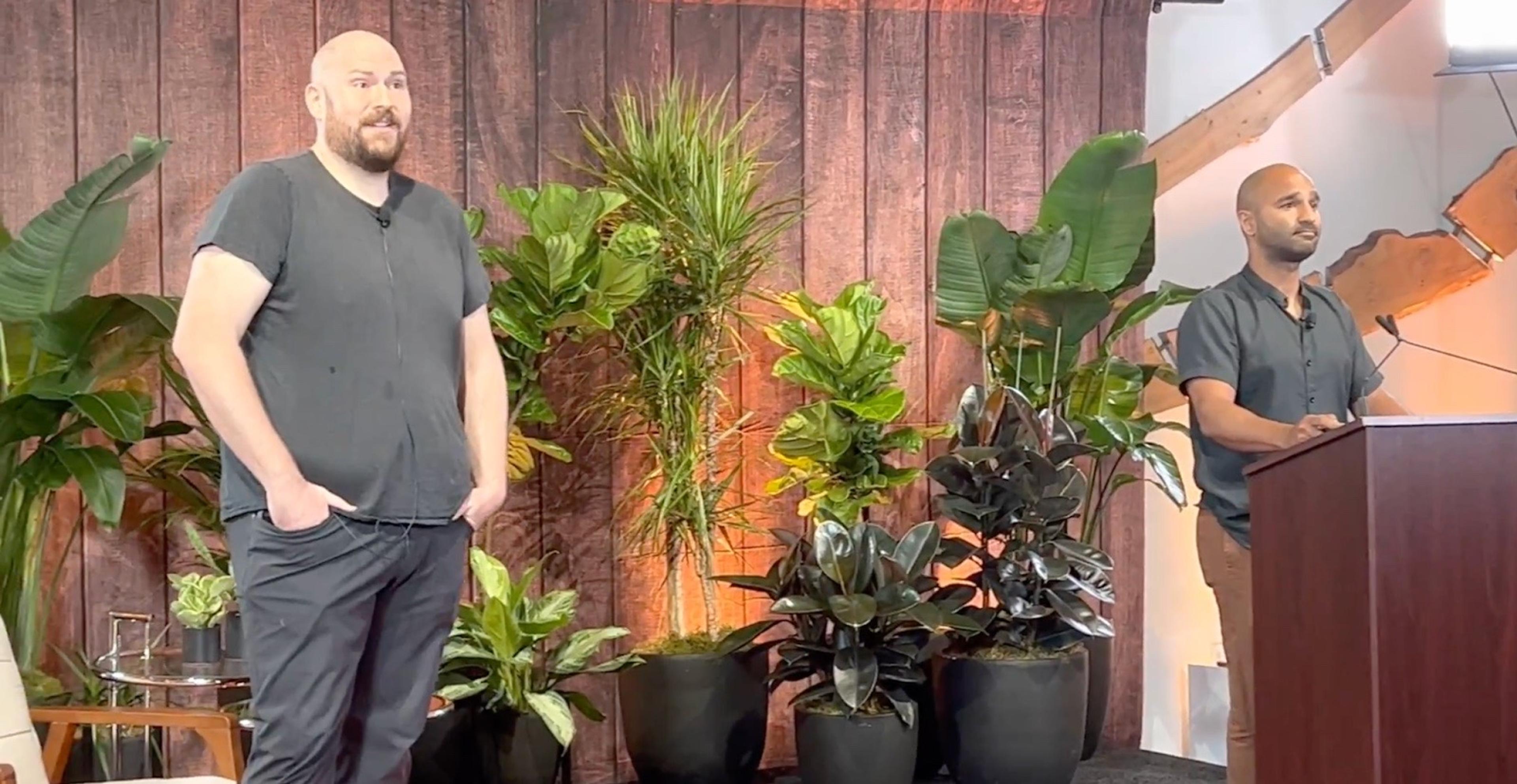 Two men with shaved heads, both wearing grey shirts, on stage presenting. One is standing with both hands in his pockets, the other is behind a podium.