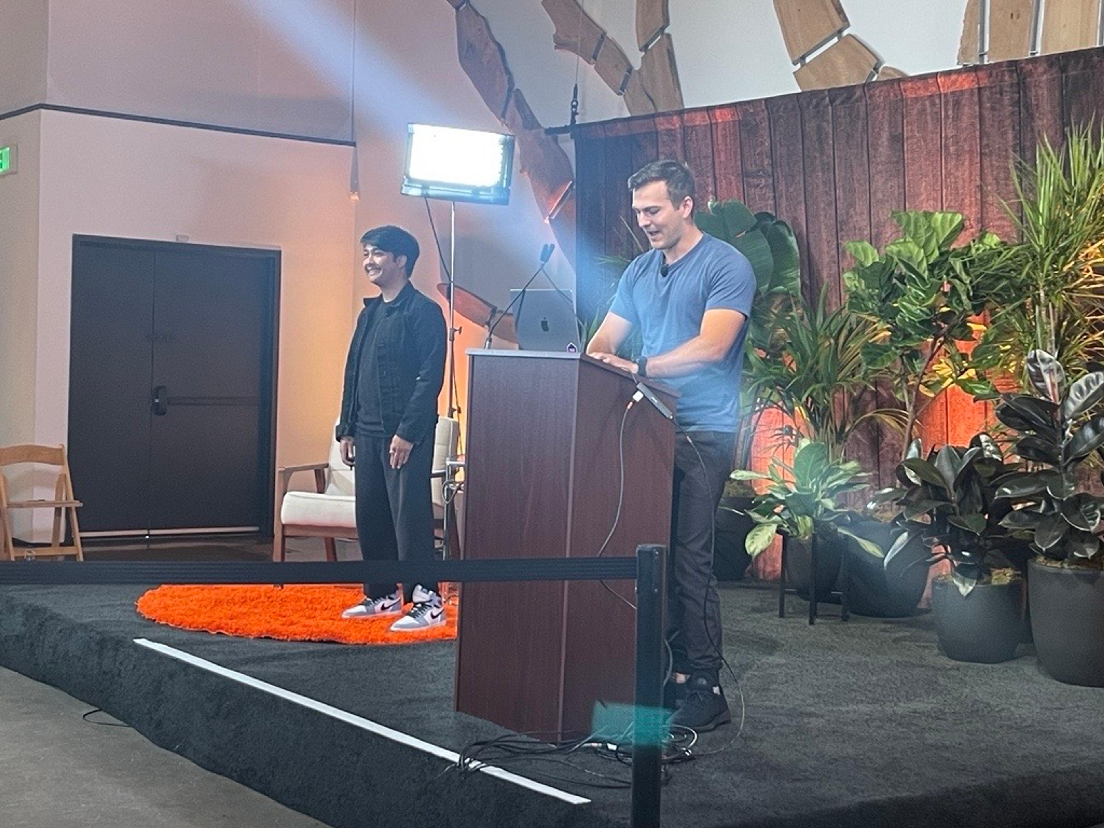 A picture of two people presenting on stage from the front-row. A man wearing all black and sneakers standing on a fuzzy orange rug and a man wearing a blue shirt and black jeans standing behind a podium.