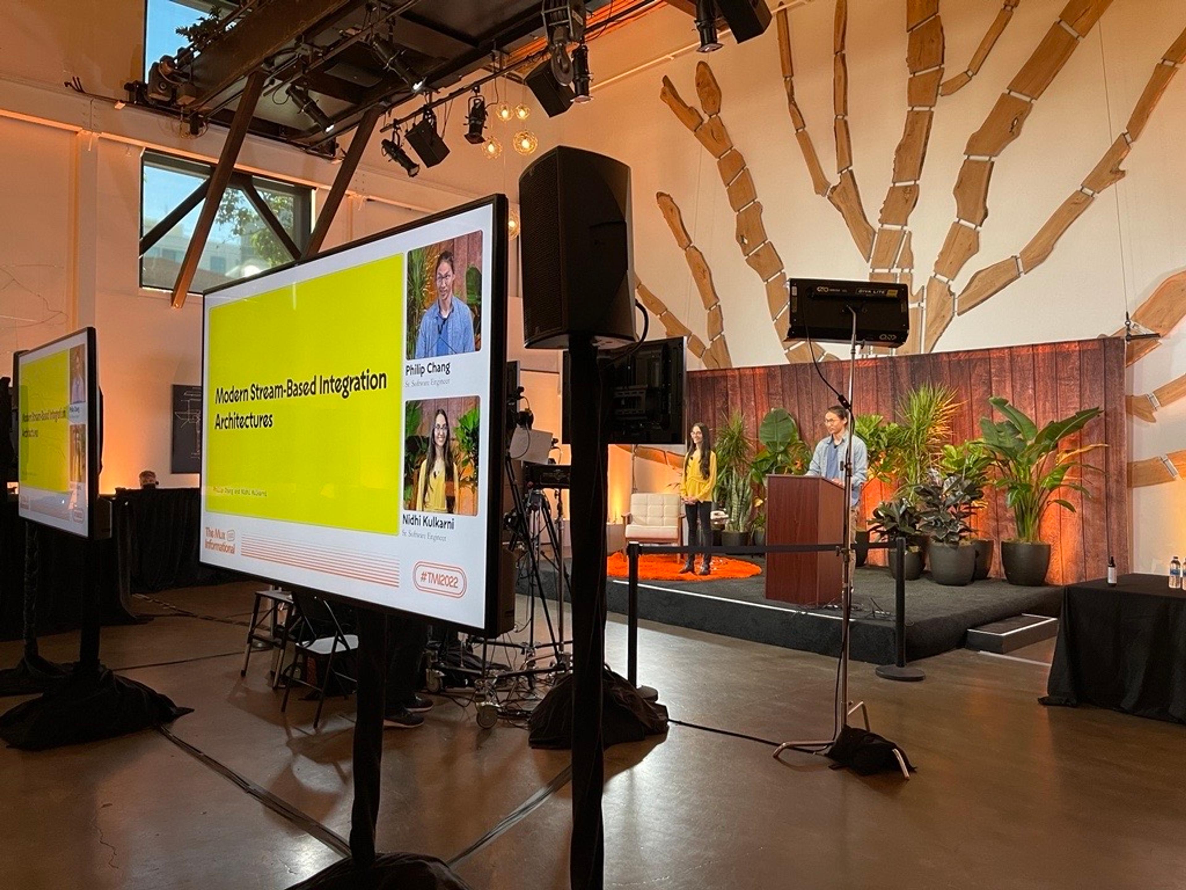 A backstage look at a man (Philip Chang)  and woman (Nidhi Kulkarni) presenting on stage. The woman wears a yellow top and hte man is wearing a light blue button down worn open with a darker blue t-shirt underneath. In the foreground, their presentation slides along with their close-ups are visible on a monitor.