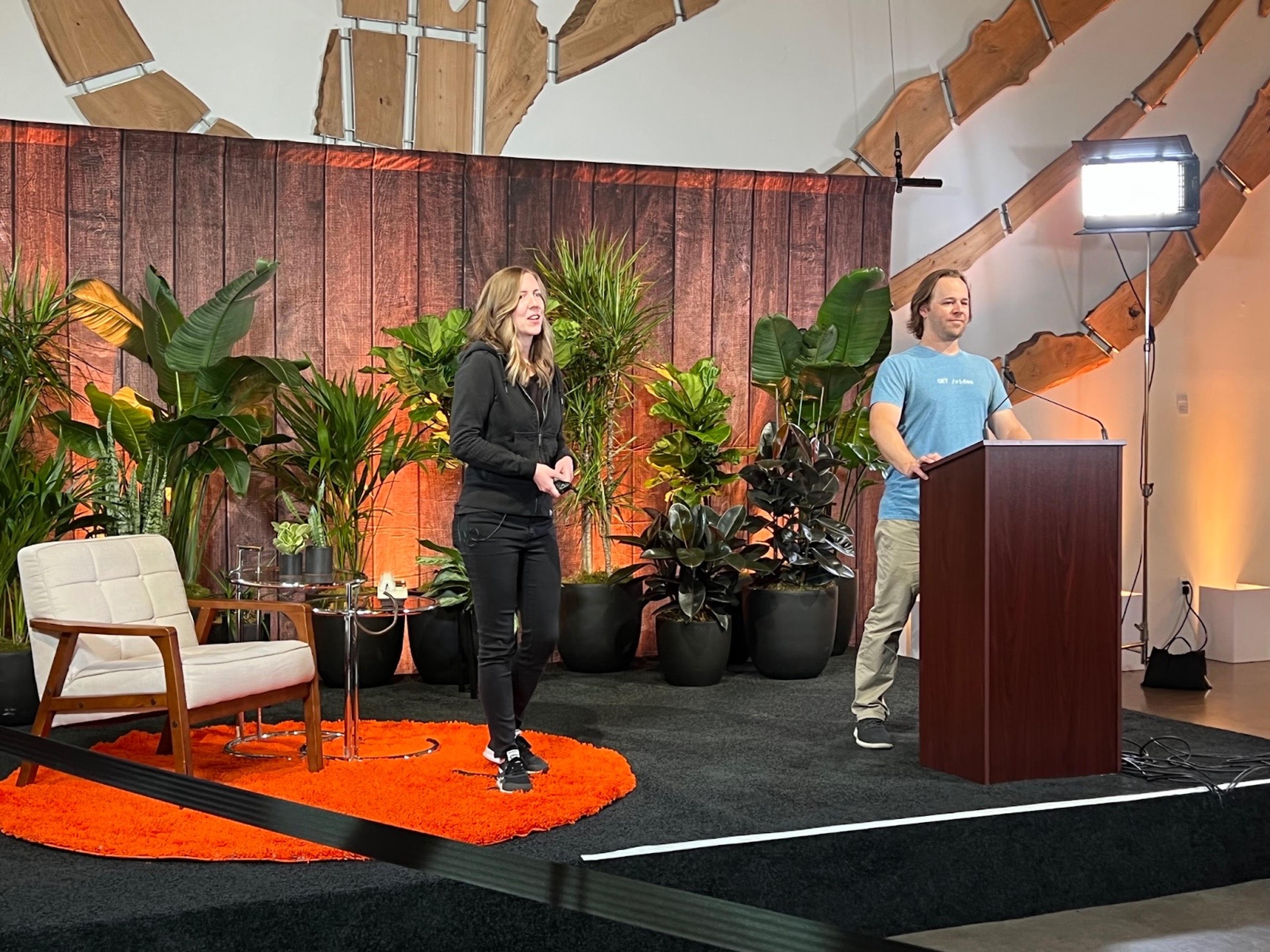 A woman with shoulder-length softly curled hair and an all-black outfit stands on stage with a man with long hair, wearing a soft blue t-shirt and beige pants. The woman stands on top of a fuzzy orange rug with a retro sitting area behind her. The man stands behind a podium.