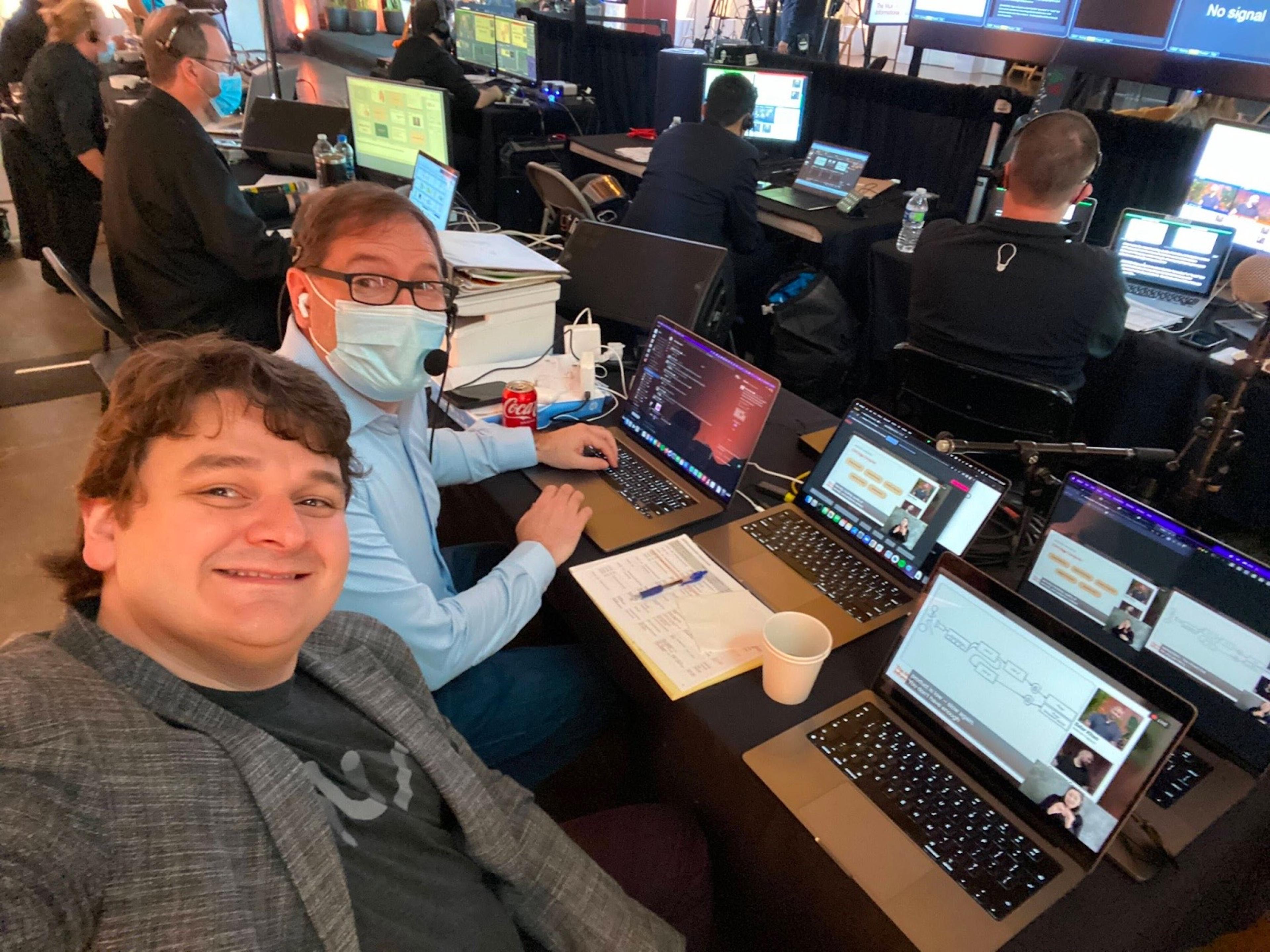 Two men taking a selfie in front of a large group of people sitting behind rows of laptop monitors.