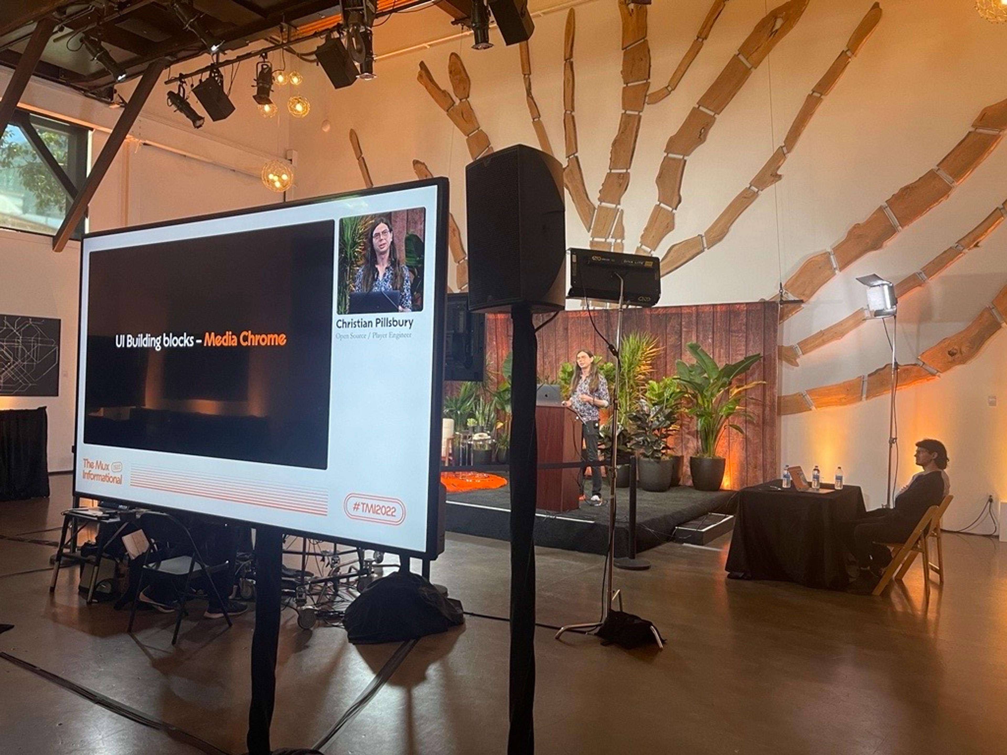 A backstage view of a man with glasses and long brown hair, wearing a colourful blue-based printed shirt presenting on stage. He is also visible on a monitor in the foreground along with his presentation slides.