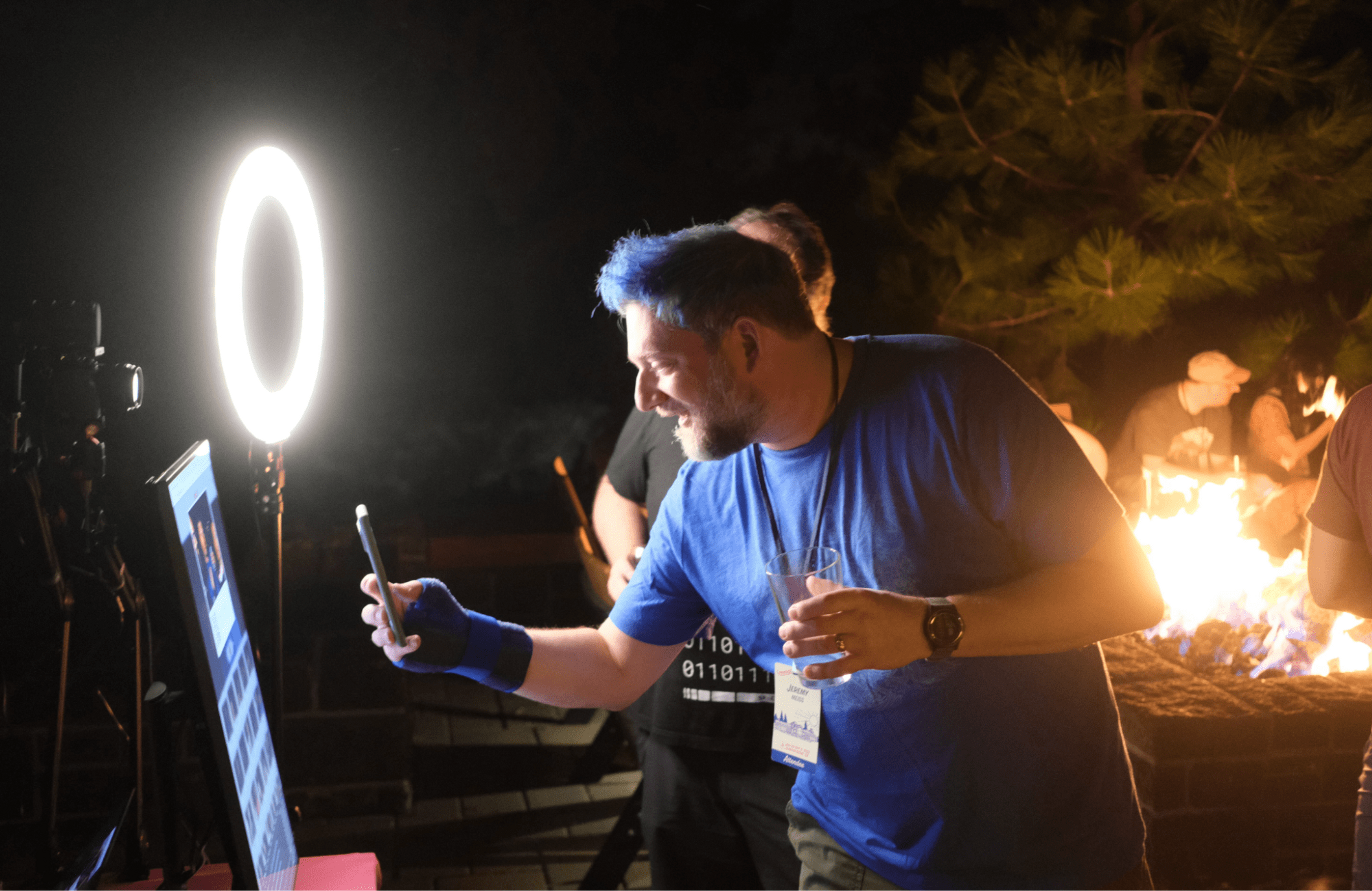 A man taking a photo of a powered-on computer monitor outside at night, next to a campfire