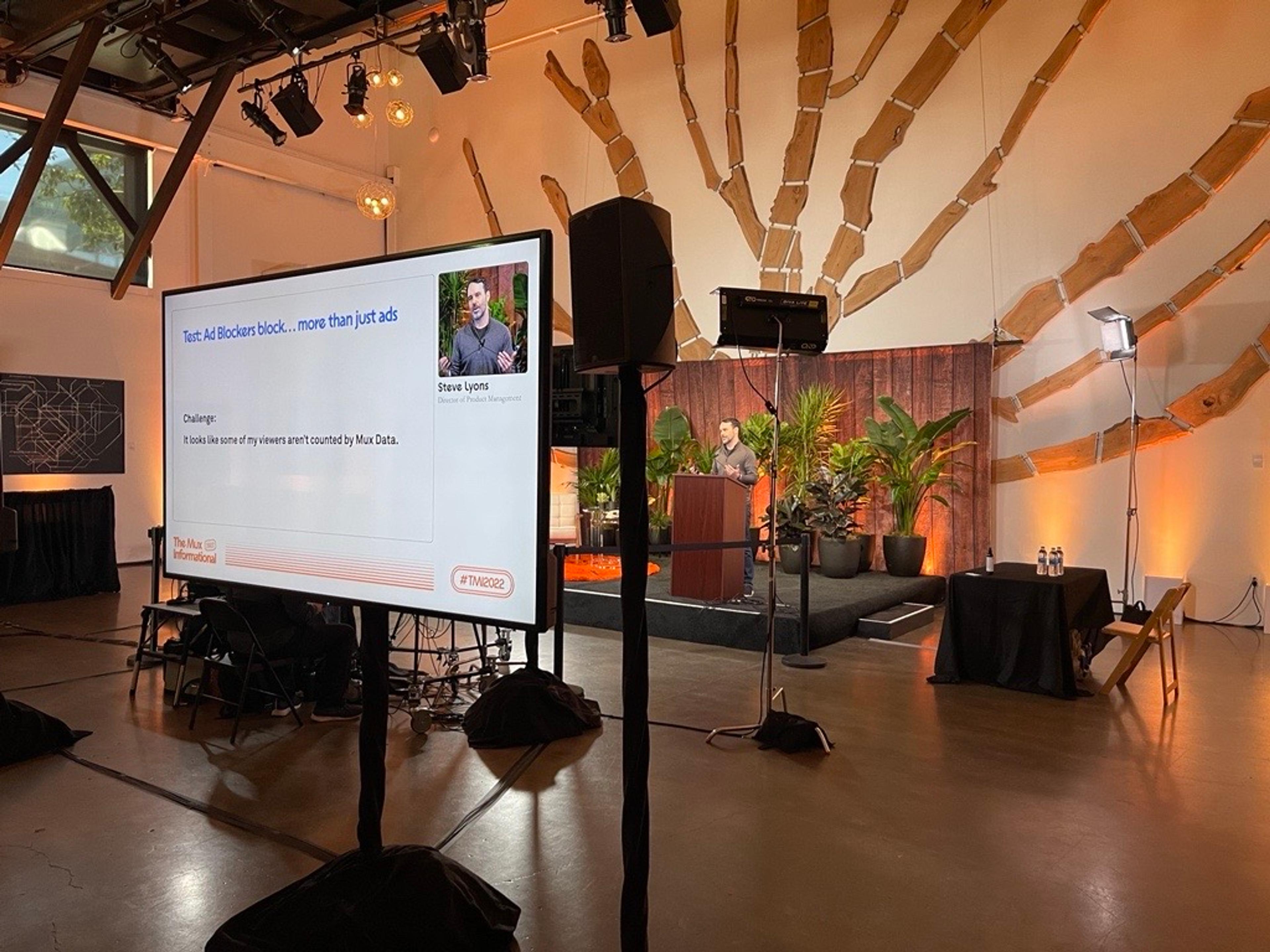 A backstage look at a man wearing a grey shirt speaking on stage behind a podium in the background. In the foreground, he is also visible with his presentation slides on a monitor.