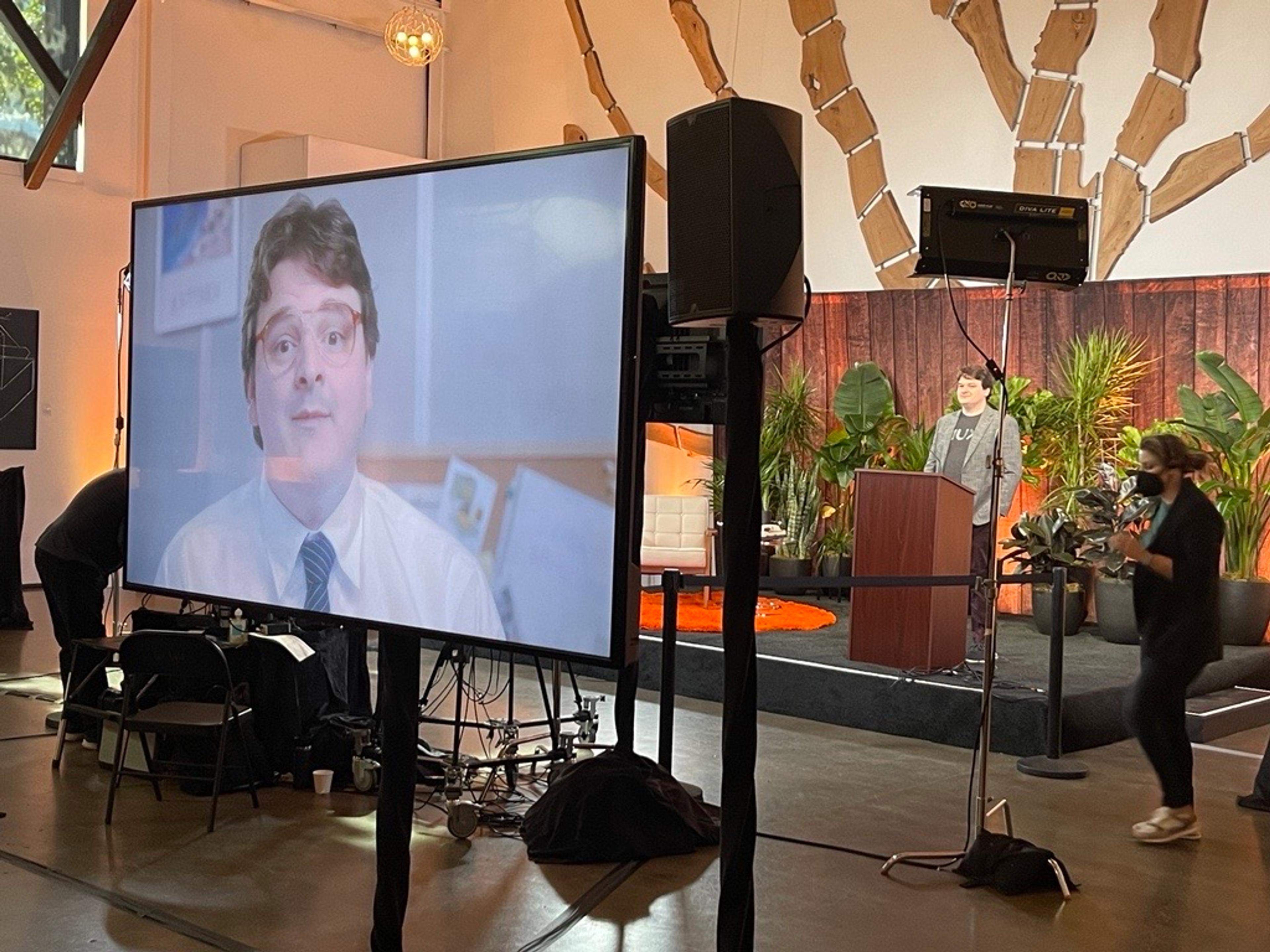 A backstage look at a man with brown hair, grey suit jacket, and black printed t-shirt presenting on stage. He is also visible on the monitor in the foreground, which displays a pre-recorded video of him in a crisp white button-up and tie.