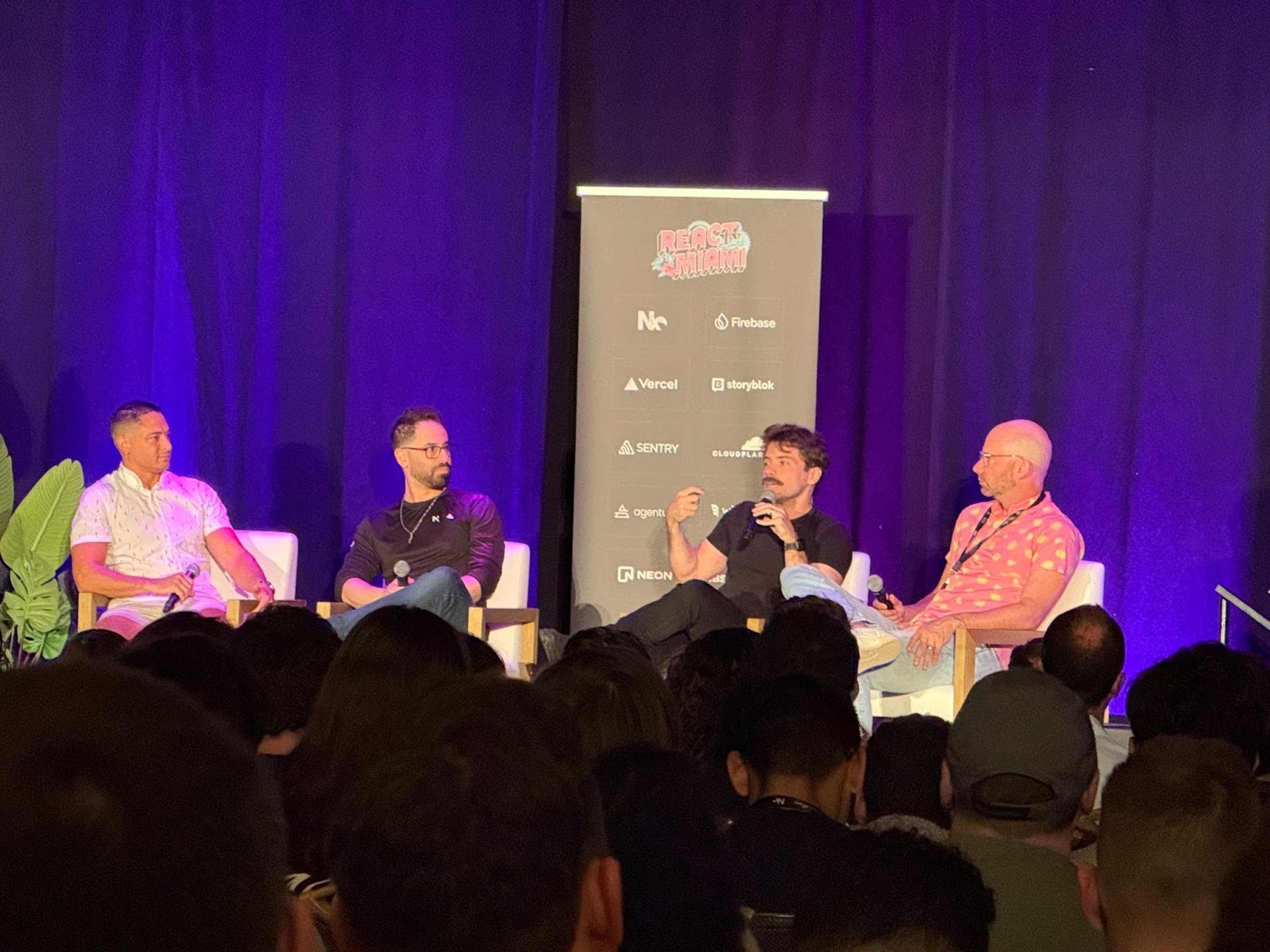 A photo of Tom Occhino, Guillermo Rauch, Taylor Otwell, and Jordan Walke, sitting and speaking in a panel setup on stage at React Miami 2025.