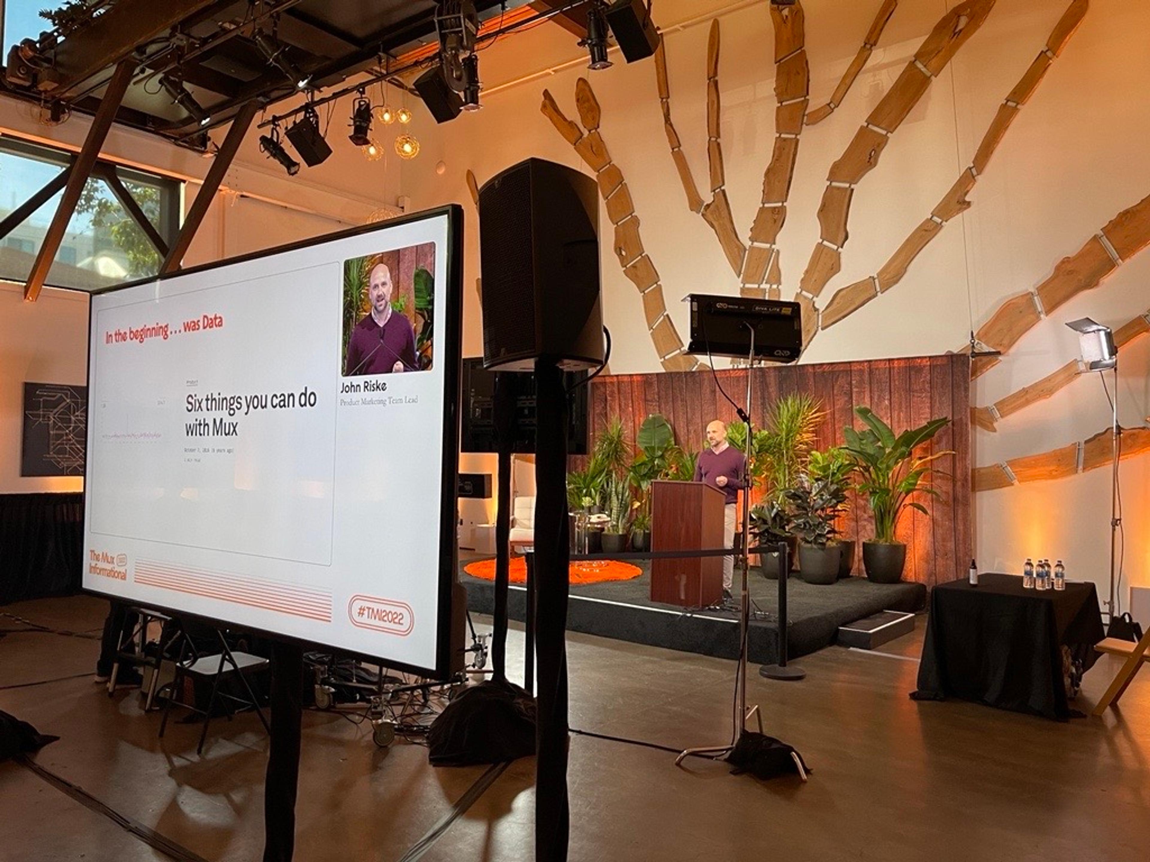 A backstage view of a man with a shaved head, wearing a burgundy shirt, named John Riske (Product Marketing Team Lead) presenting on stage. In the foreground, him and his presentation slides saying, "Six things you can do with Mux," are visible on a monitor.
