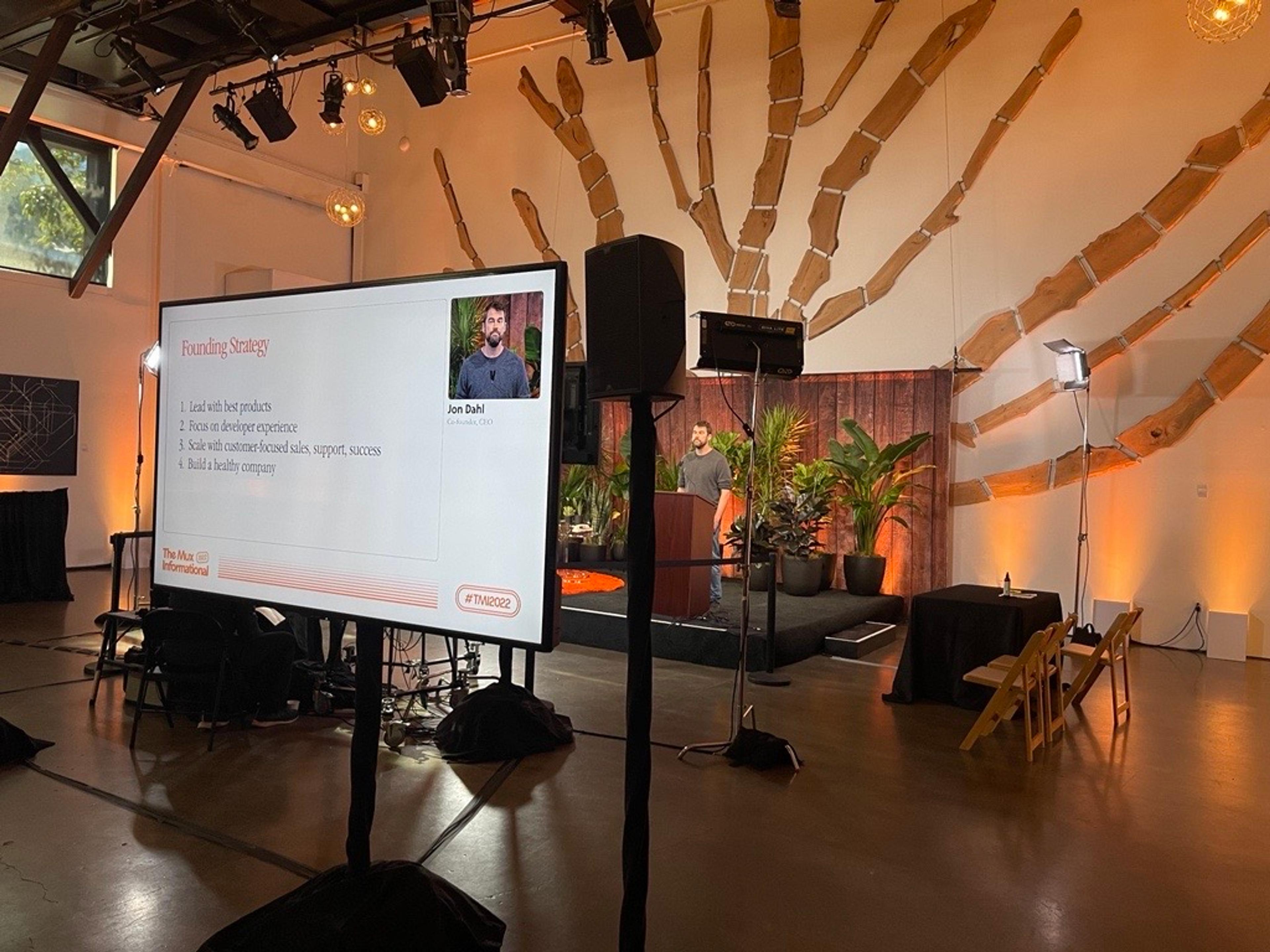 Behind the stage picture of a man in a grey shirt speaking on stage. He is also visible in the foreground with his presentation slides on a monitor.