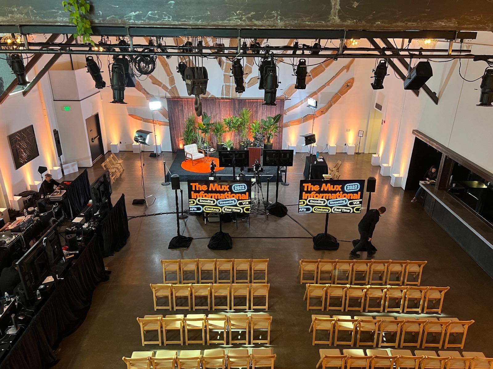 Overhead view of a venue space. On the ground floor, a stage is visible with large monitors in front, multiple rows of wooden chairs in the foreground. Off to the left, long black tables with video monitoring equipment. Overhead lights are visible.