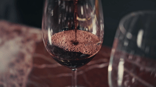 Red wine being poured into a wine glass in slow motion, with visible bubbles and foam forming at the surface. A second empty wine glass is partially visible in the background. The scene is shot on a dark, moody background with warm, dramatic lighting.