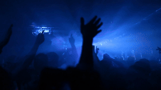 A slow-motion GIF of a crowd at a concert or club event, shot from behind. Hands are raised against a backdrop of dramatic blue stage lighting with light beams cutting through haze above the crowd.