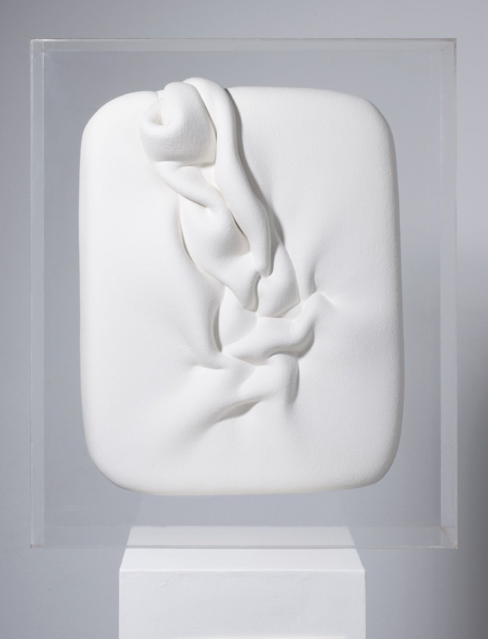Ein weißes Schaumstoffobjekt in Grundform eines abgerundeten Rechtecks. Der Schaumstoff ist auf der Vorderseite in Falten gelegt, die deutlich hervorstehen. Das Objekt befindet sich in einem Acrylglas-Kasten.
