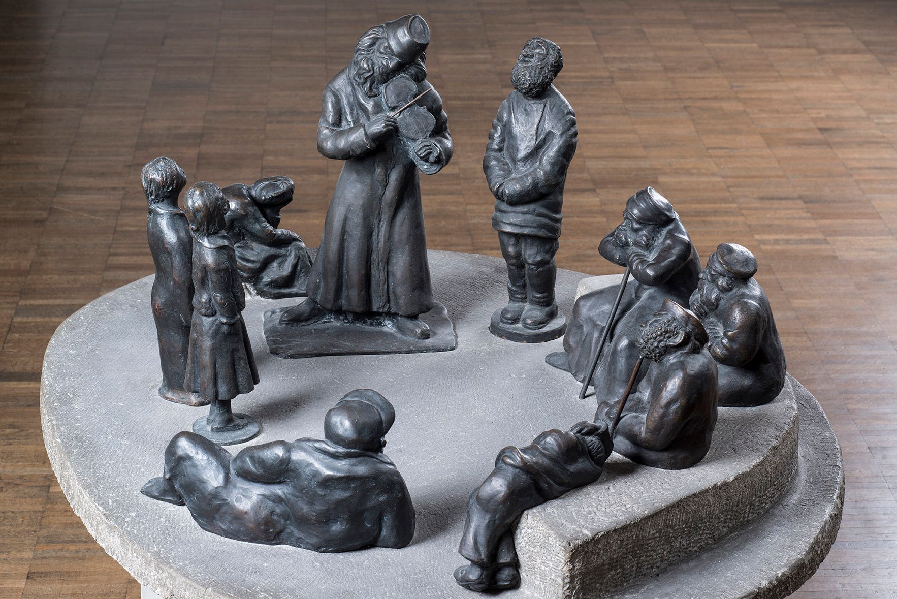 Skulpturengruppe von 10 Figuren auf einer runden Steinplatte. Ein Mann spielt die Geige, die anderen hören ihm andächtig zu. Drei alte Bärtige Männer mit Gehstöcken sitzen auf einer Bank. Mann stützt sich auf dem Boden liegend mit einem Arm ab, ein Junge und ein Mädchen hören zu.