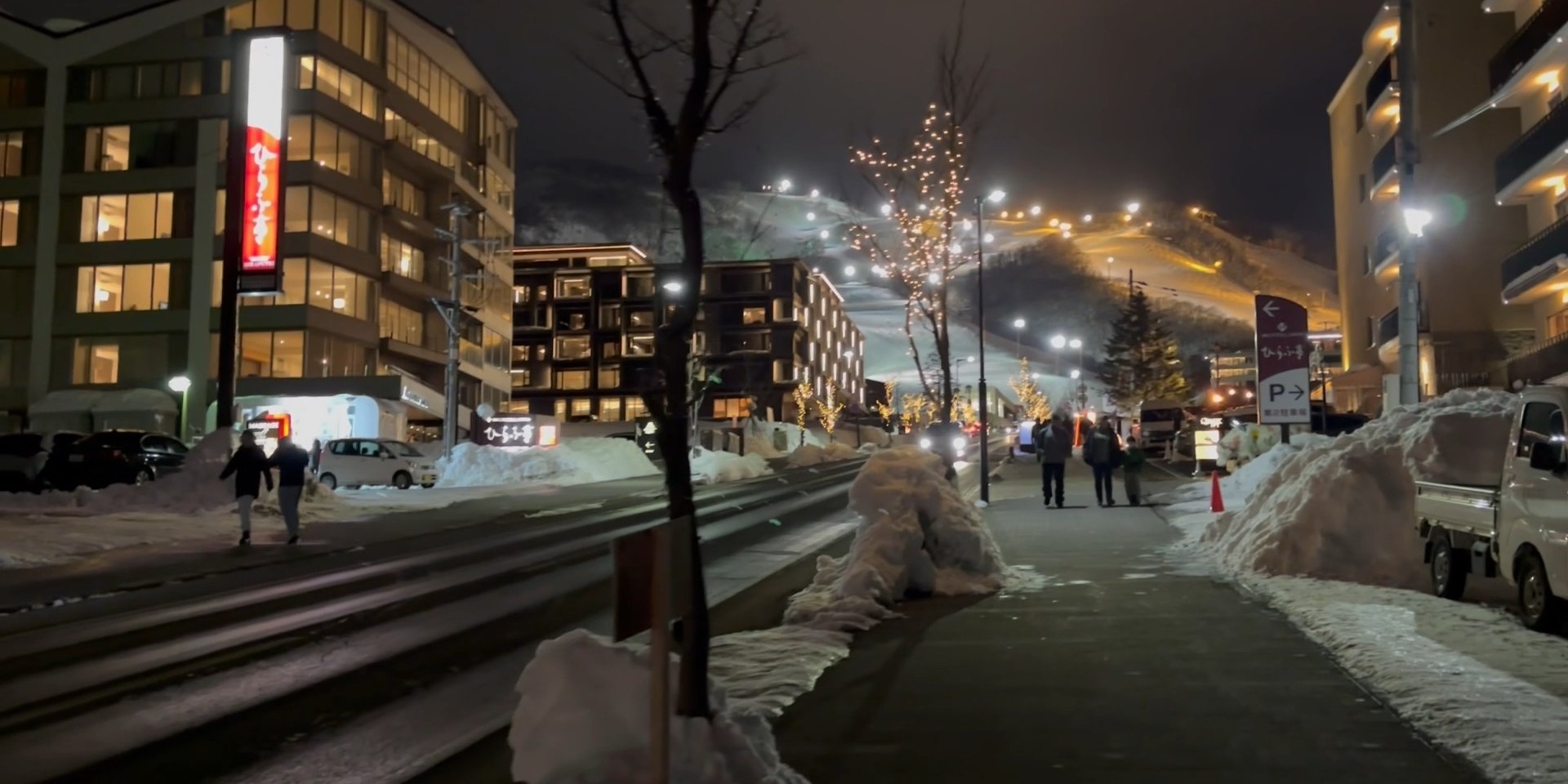 Fairy lights and lit shopfronts along a snowy Niseko village street at night
