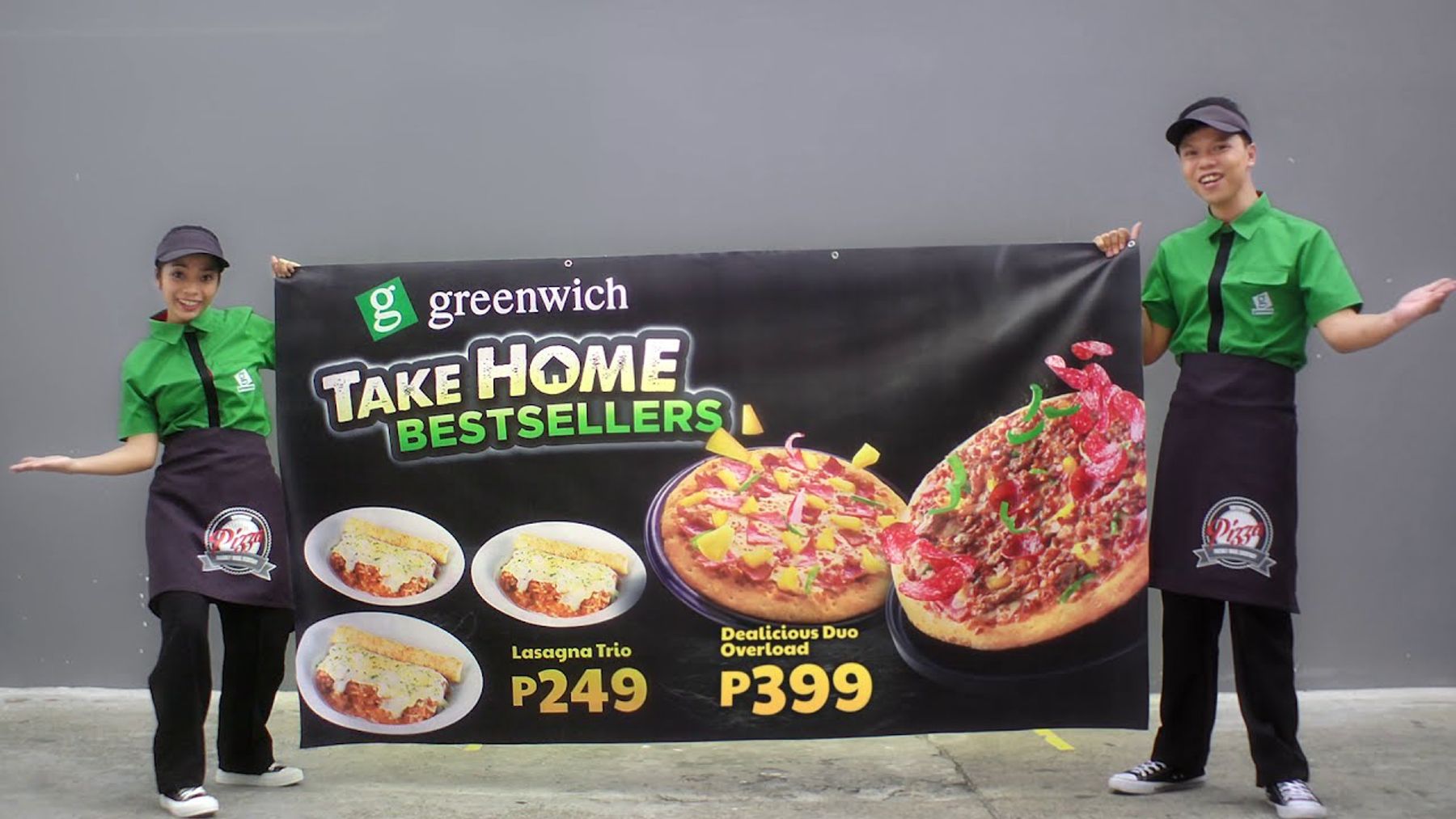 a man and a woman dressed in green hold up a black banner with photos of pizza and lasagna on it