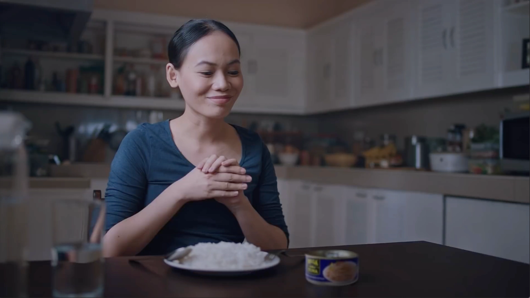 A woman in a kitchen looks at a can of Mega Tuna. Her plate is full of rice and she is ready to eat 