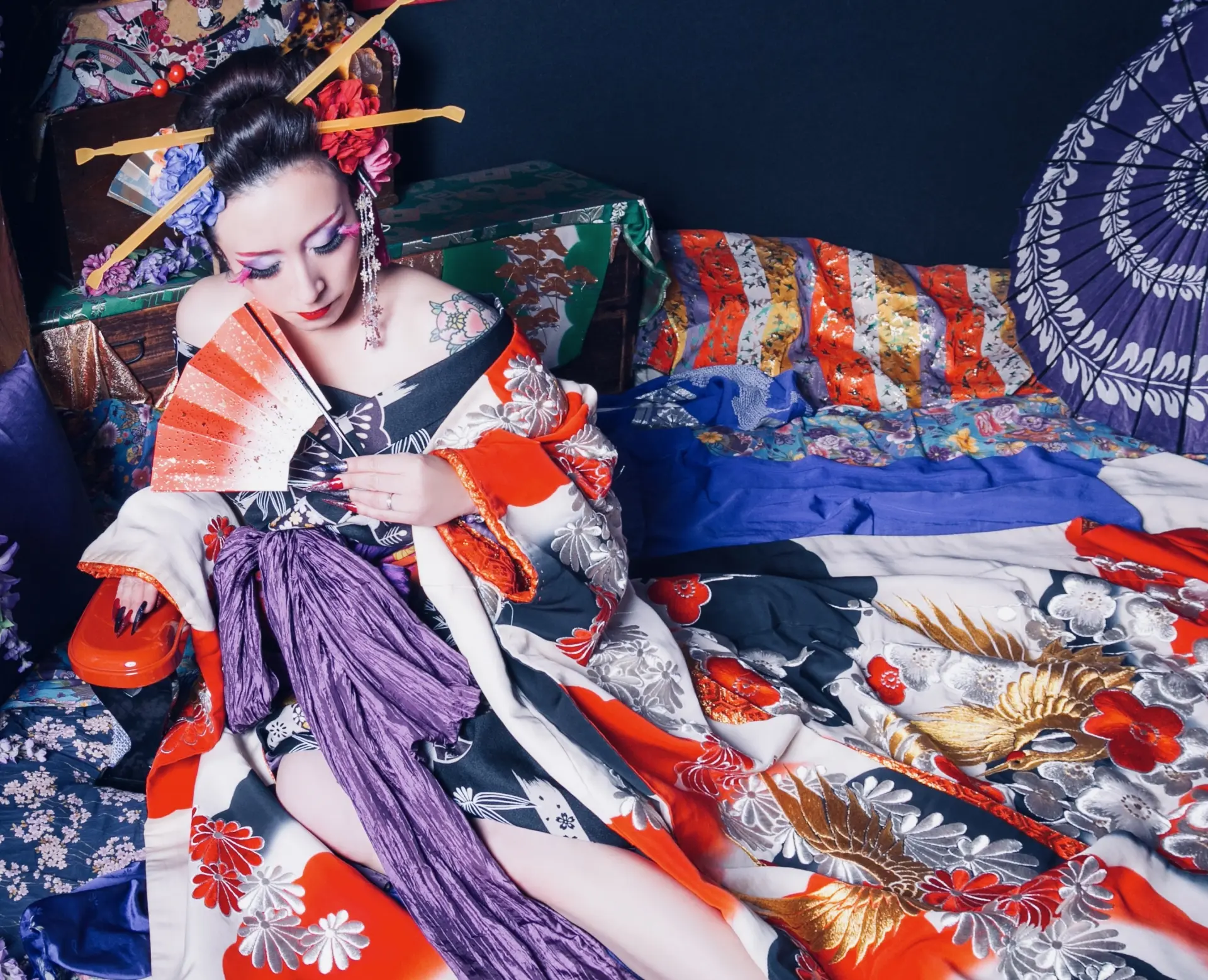 A woman in an ornate, kimono with dramatic makeup and decorated hair, holding a fan while reclining amidst colorful fabrics.