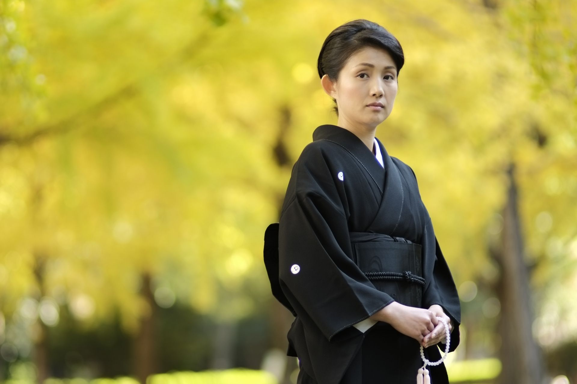 Japanese woman in a black kimono holding prayer beads, with yellow ginkgo trees in the background.