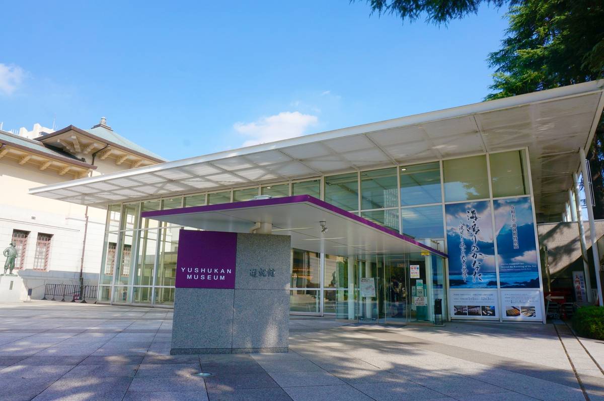 The modern glass-fronted entrance of the Yushukan Museum with a purple sign, under a bright blue sky.