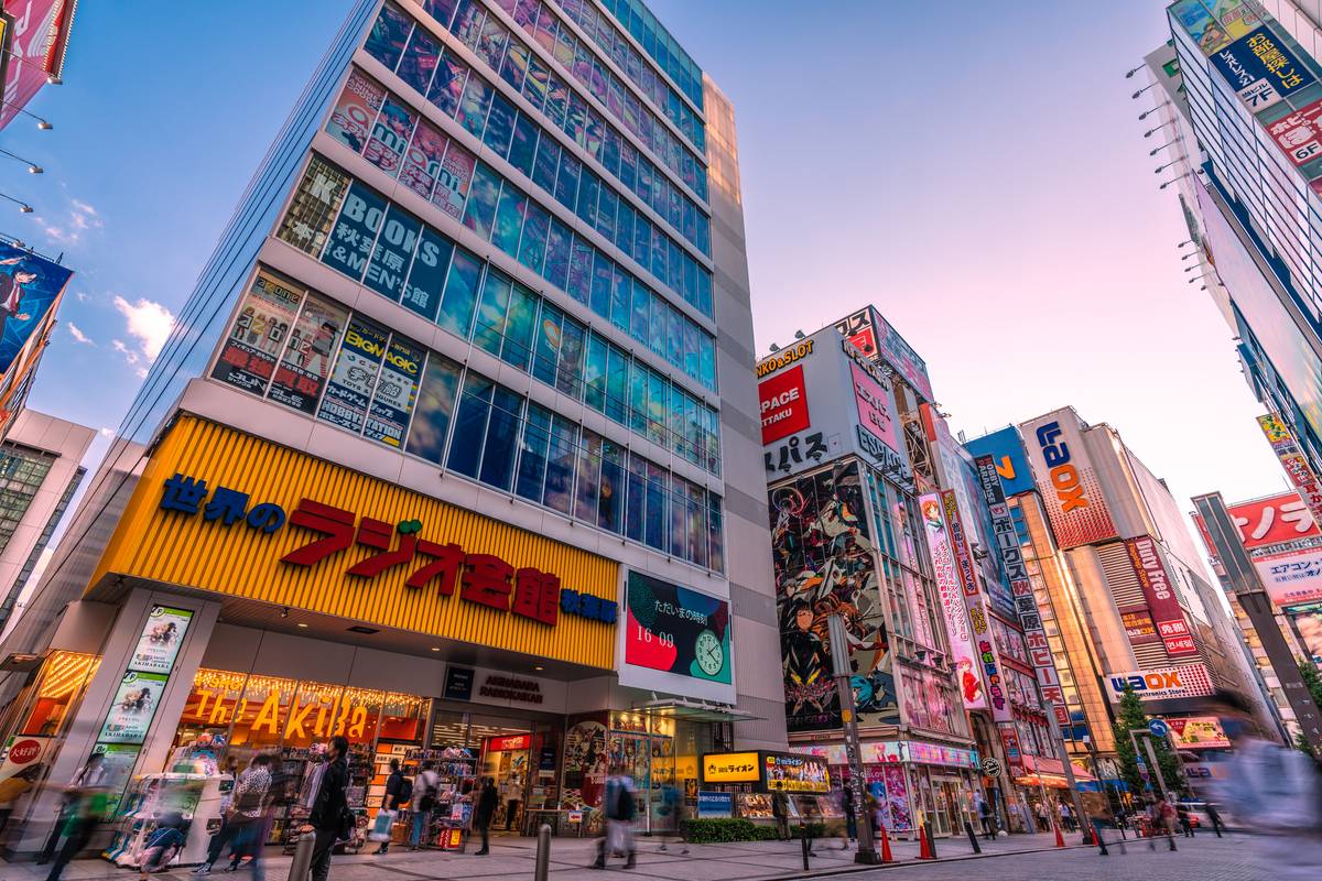 Bustling Akihabara street with tall buildings covered in colorful Japanese signs under a blue and pink sky.