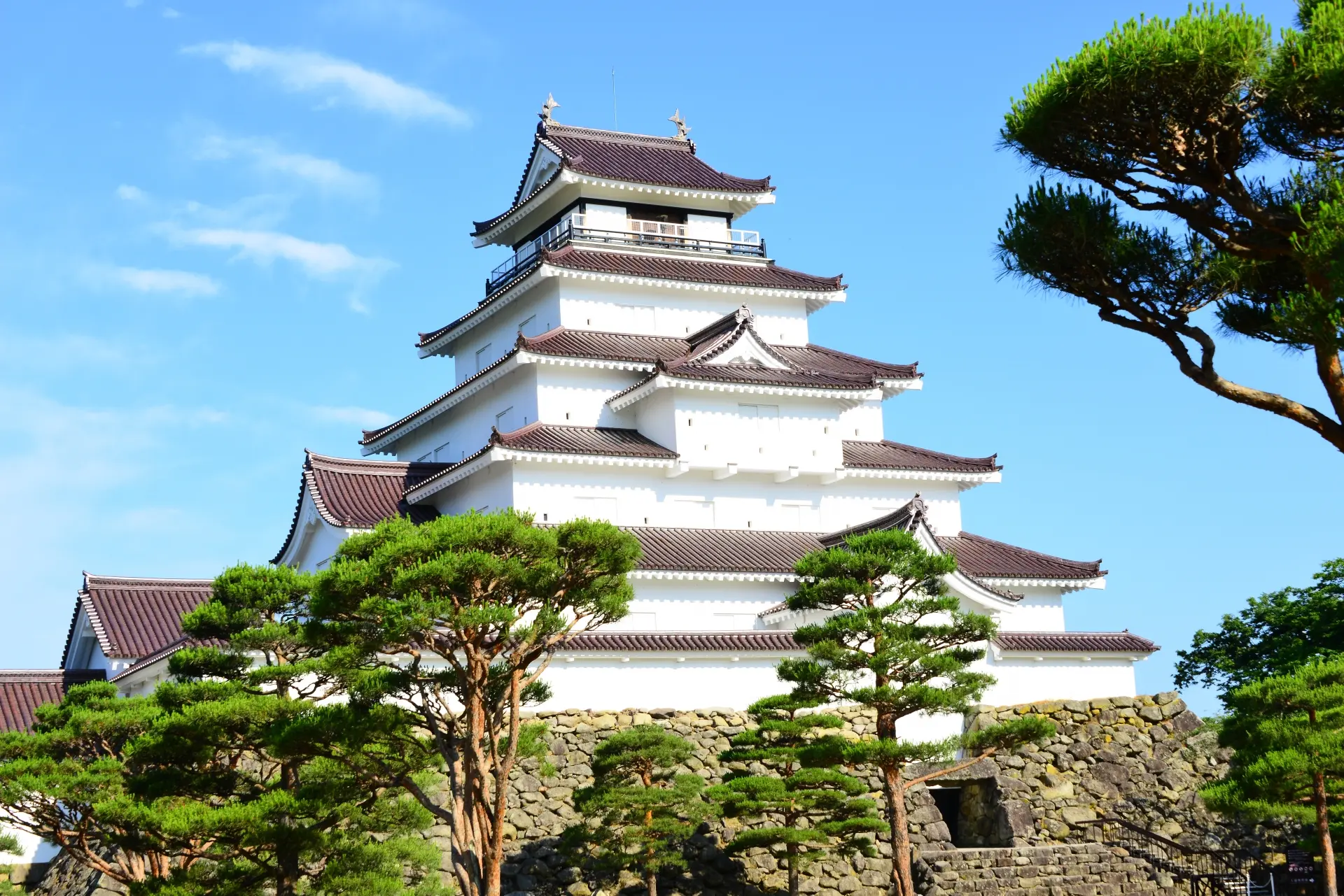 A white multi-tiered Japanese castle with dark roofs, surrounded by green pine trees, under a blue sky.