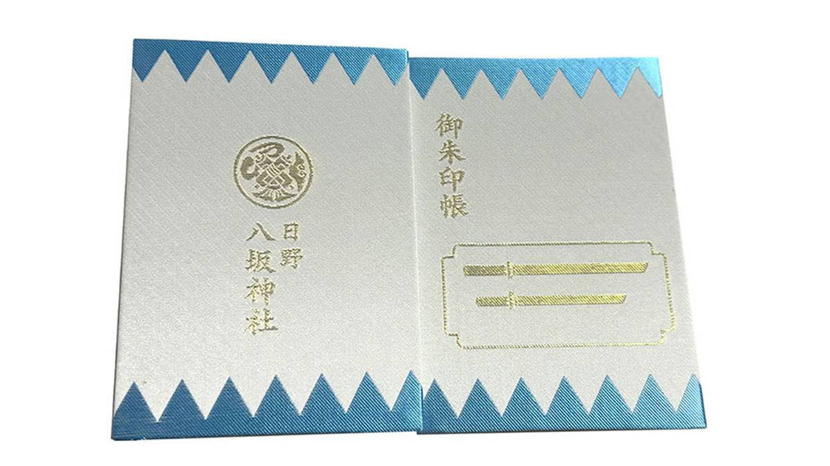 An open Japanese Goshuincho with white pages, blue zigzag borders, gold Japanese text, and sword illustrations.