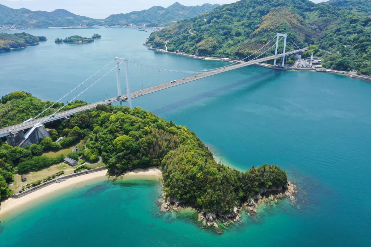 Aerial view of a suspension bridge over turquoise water connecting green, mountainous land, with a sandy beach.