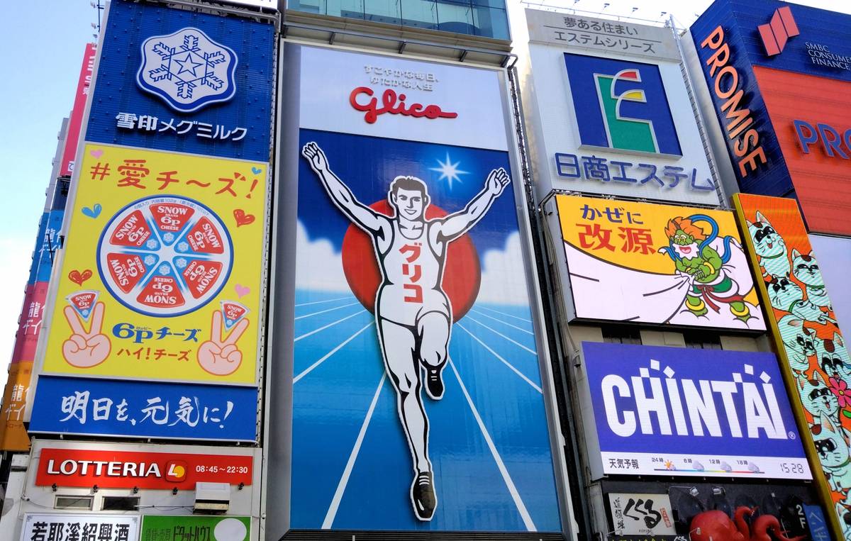 A building facade covered in numerous vibrant Japanese advertisements, dominated by the large Glico Man billboard in the center.