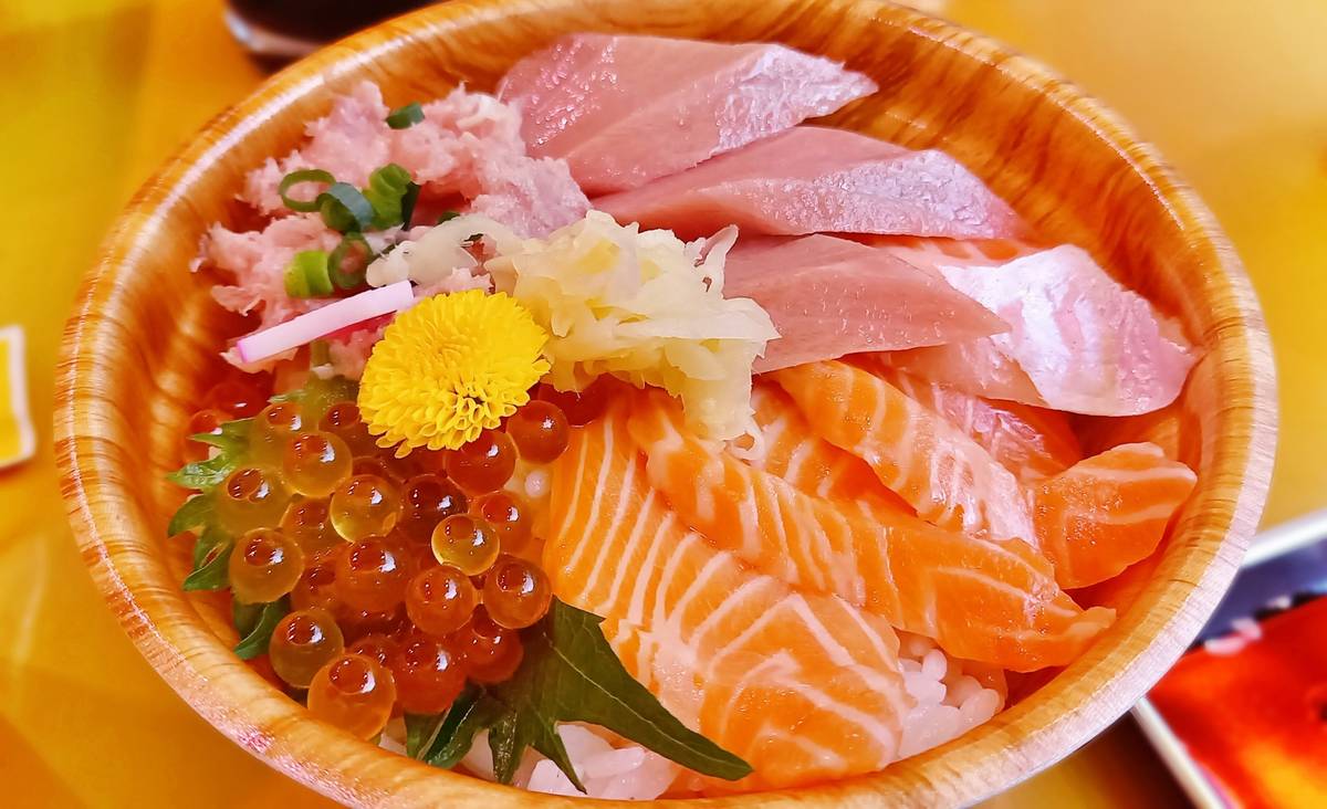 Chirashi sushi bowl with salmon, tuna, minced fish, and salmon roe, garnished with ginger and a yellow flower.