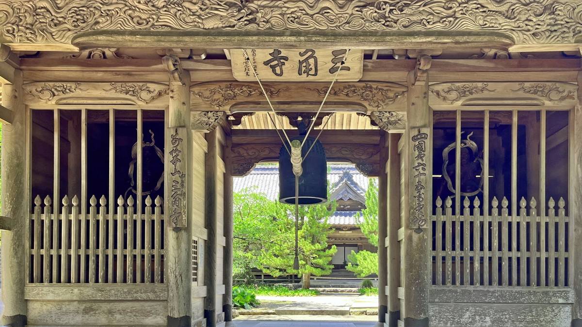 Intricately carved wooden temple gate with a large hanging bell, opening to a view of temple grounds and trees.