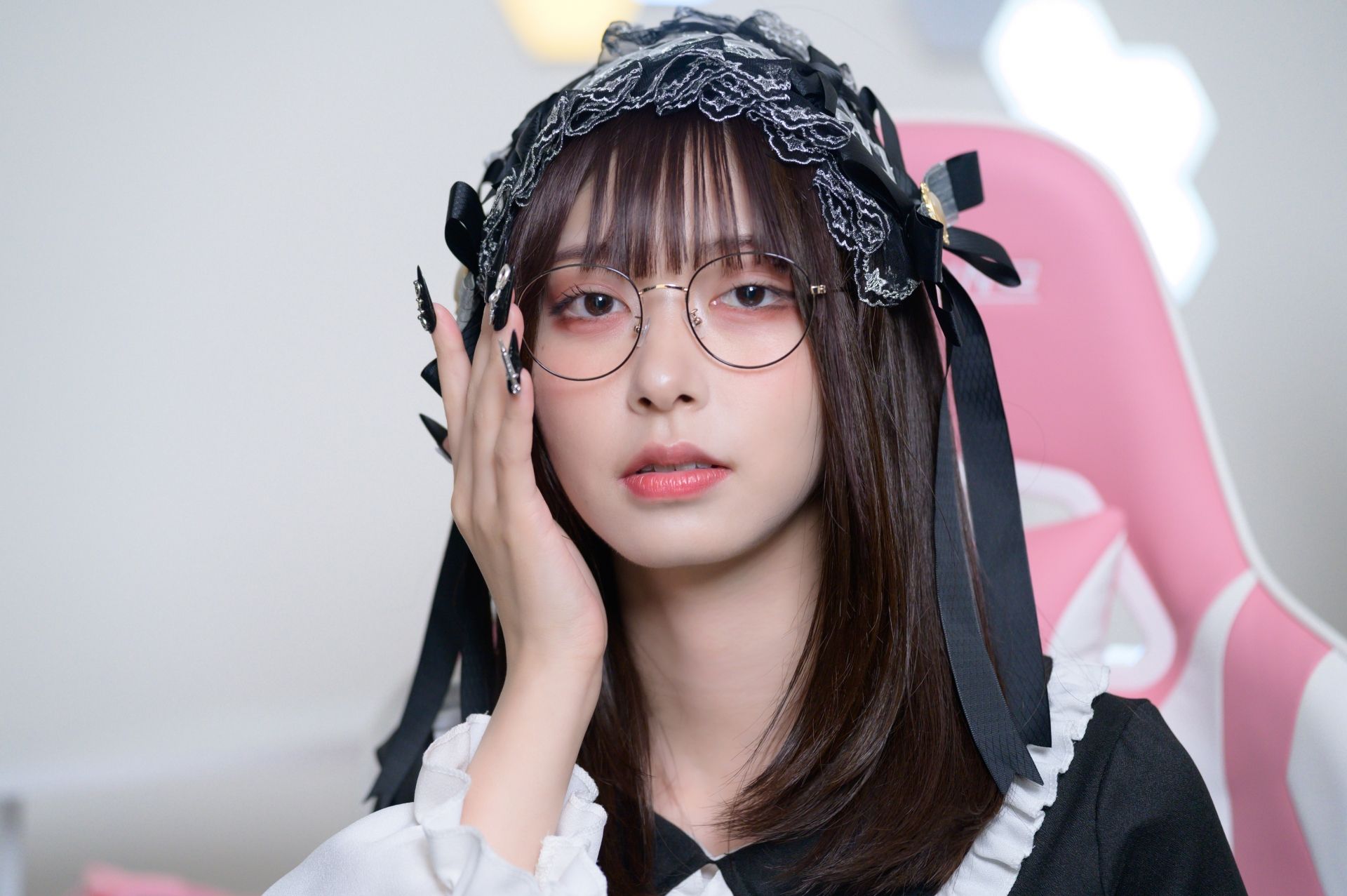 A young woman with bangs, round glasses, and long dark nails wears a black lace headband and a maid-style outfit