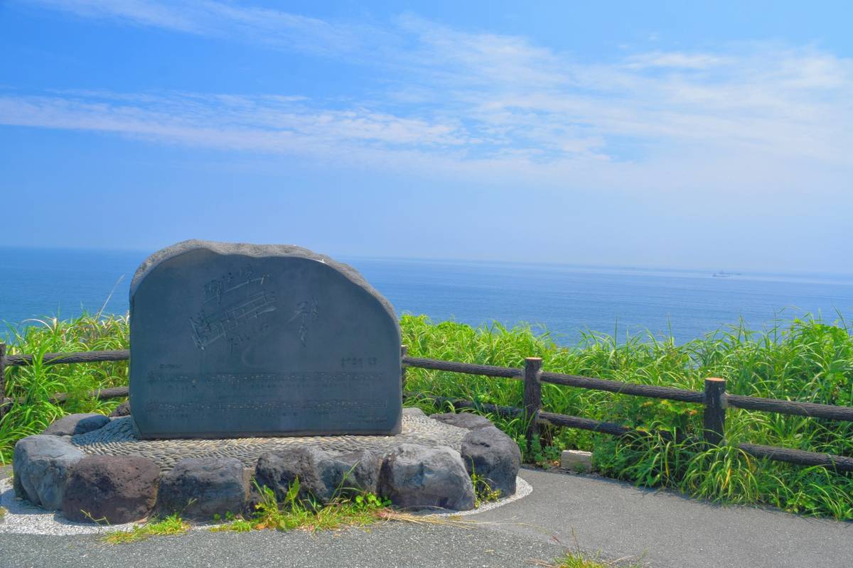 A dark stone monument on a cliff overlooking a vast blue ocean under a clear sky, with green grass and a wooden fence.