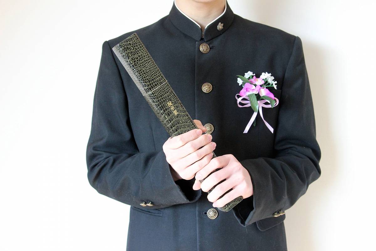 A student in a black uniform with a corsage holds a textured diploma case.
