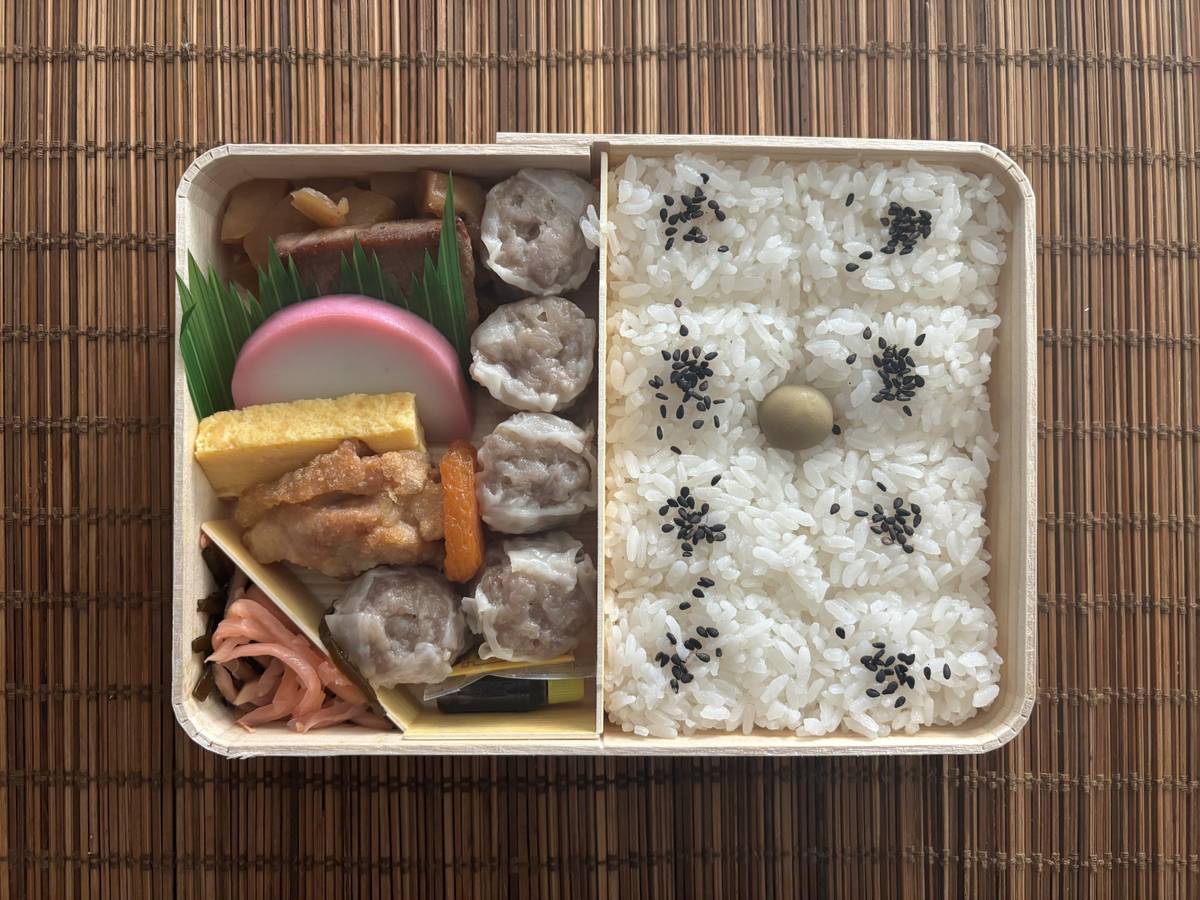 A bento box with rice, shumai, tamagoyaki, fish cake, and other Japanese side dishes on a bamboo mat.