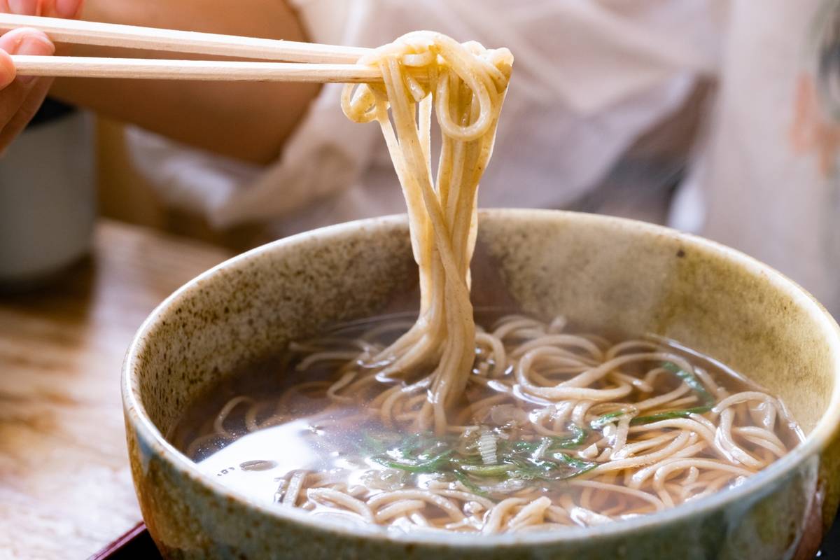 A hand uses chopsticks to lift soba noodles from a bowl of hot broth.