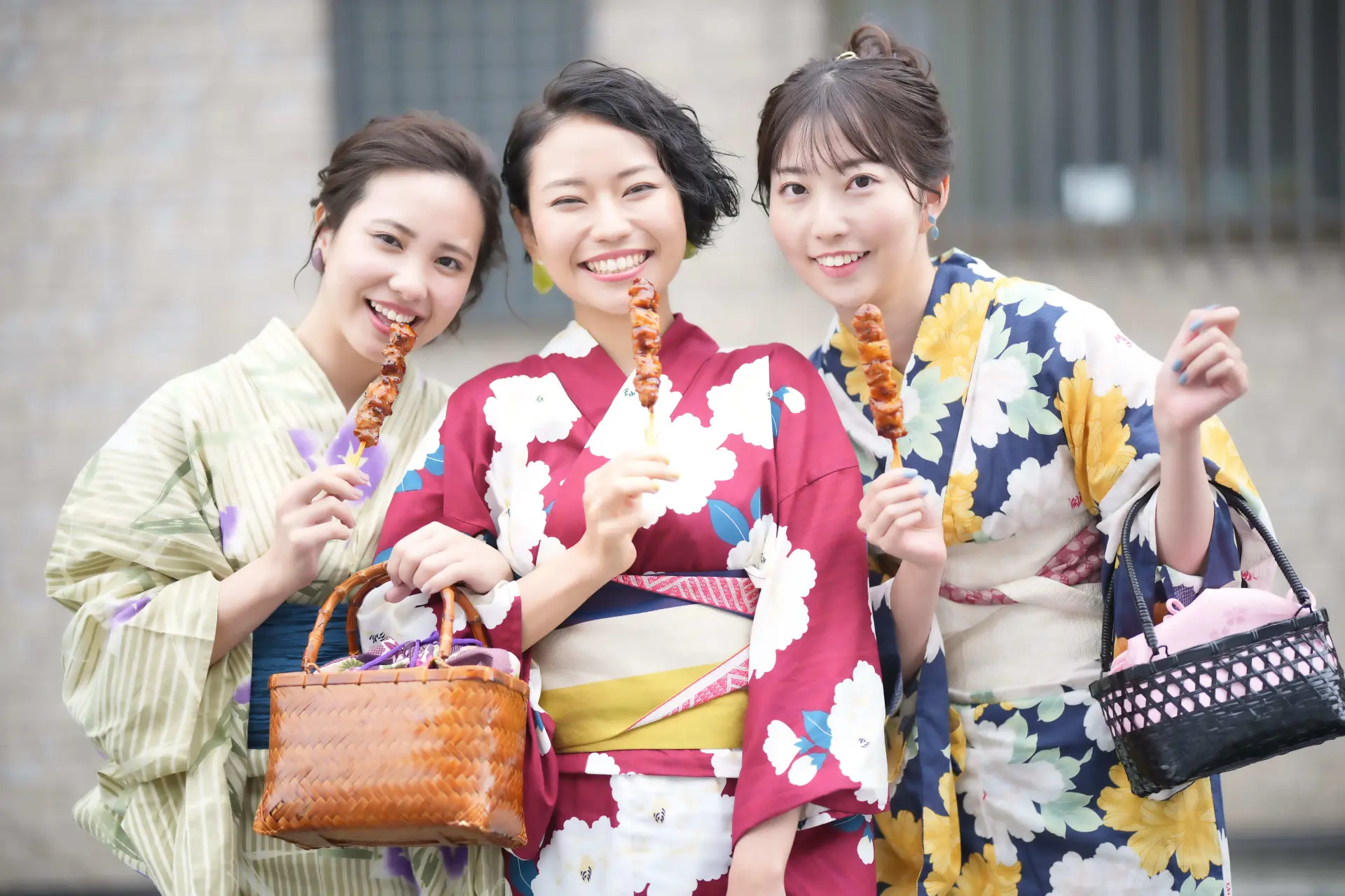 Three women in colorful yukata smile, holding skewers and traditional bags.