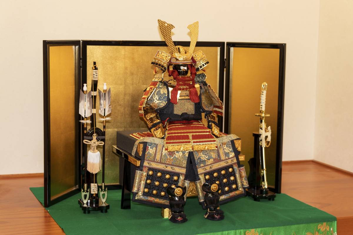 An elaborate samurai doll in full armor, displayed on a green mat in front of a gold screen, flanked by ceremonial arrows and a katana.