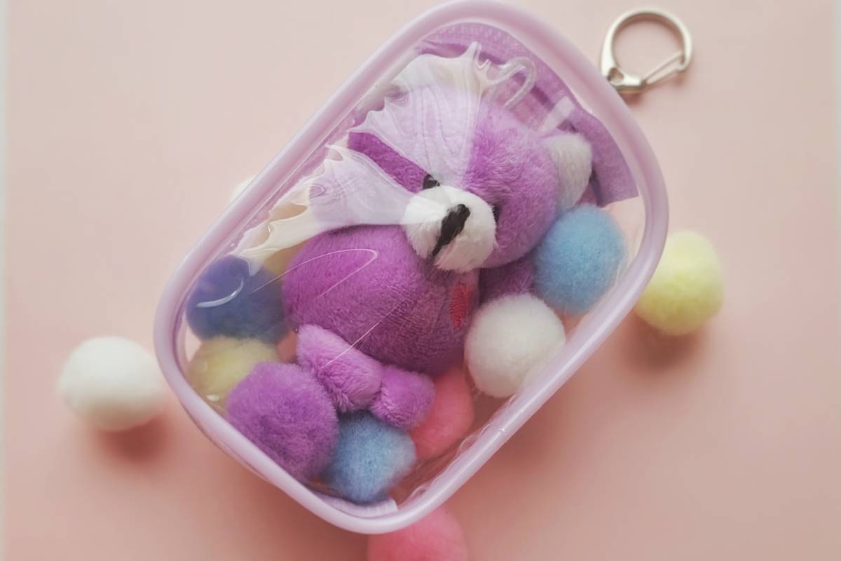 A clear pouch keychain containing a purple teddy bear and colorful pom-poms, on a pink surface with scattered pom-poms.