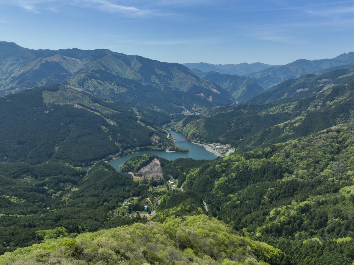 Aerial view of lush green mountains surrounding a winding lake under a blue sky.