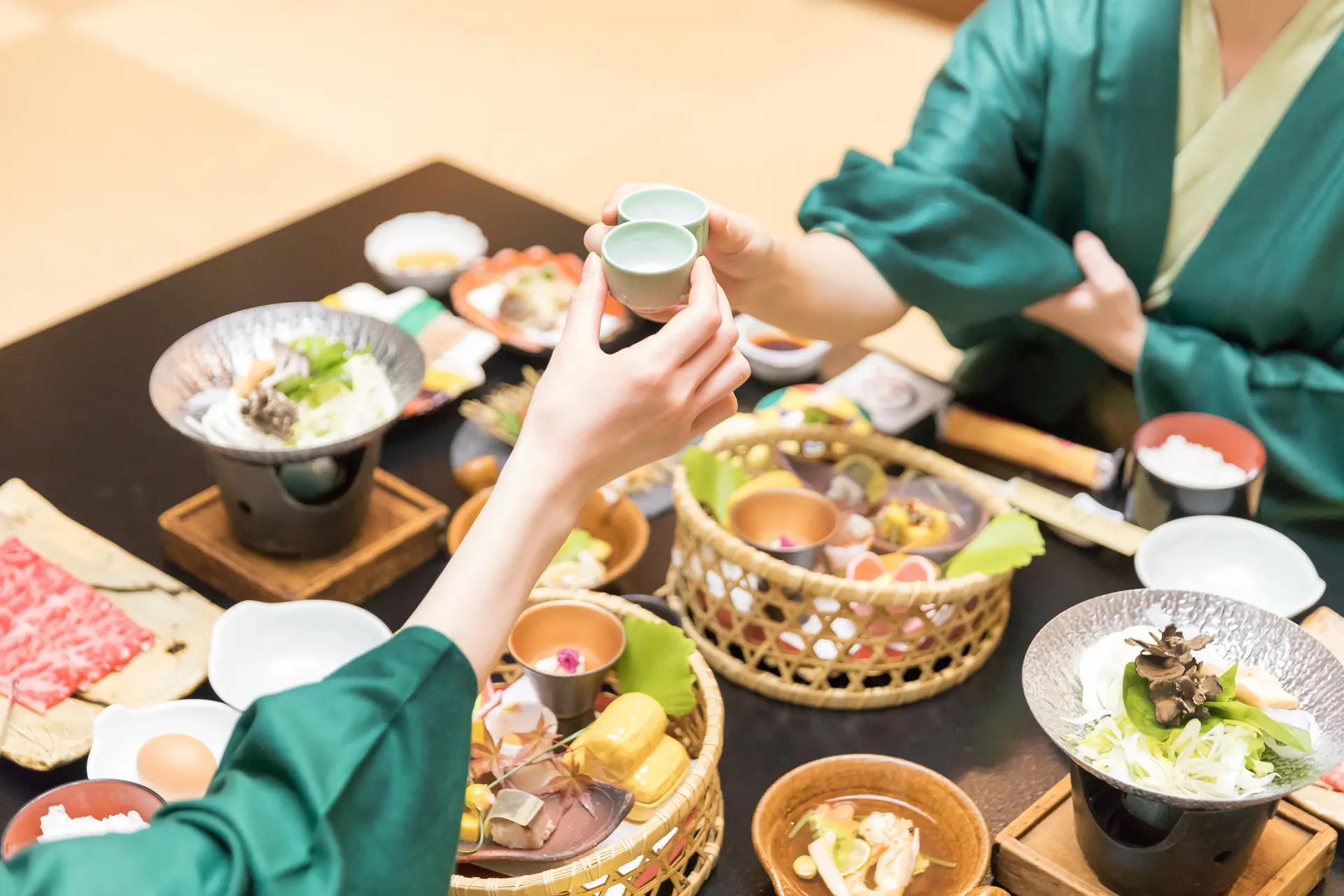 Two people in green robes clinking small ceramic cups during a traditional Japanese meal.