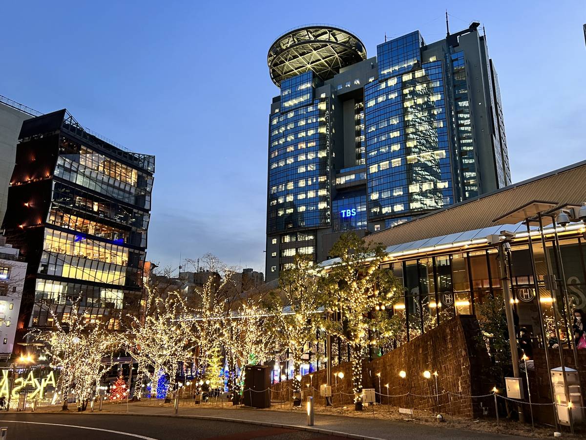 Modern city street scene at dusk with buildings and trees illuminated by string lights.