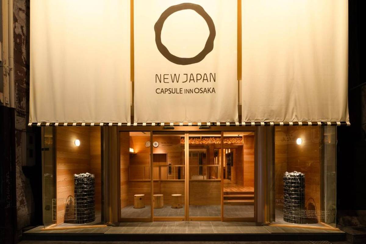 Illuminated entrance of New Japan Capsule Inn Osaka at night, with a sign and stone-filled columns visible through the windows.