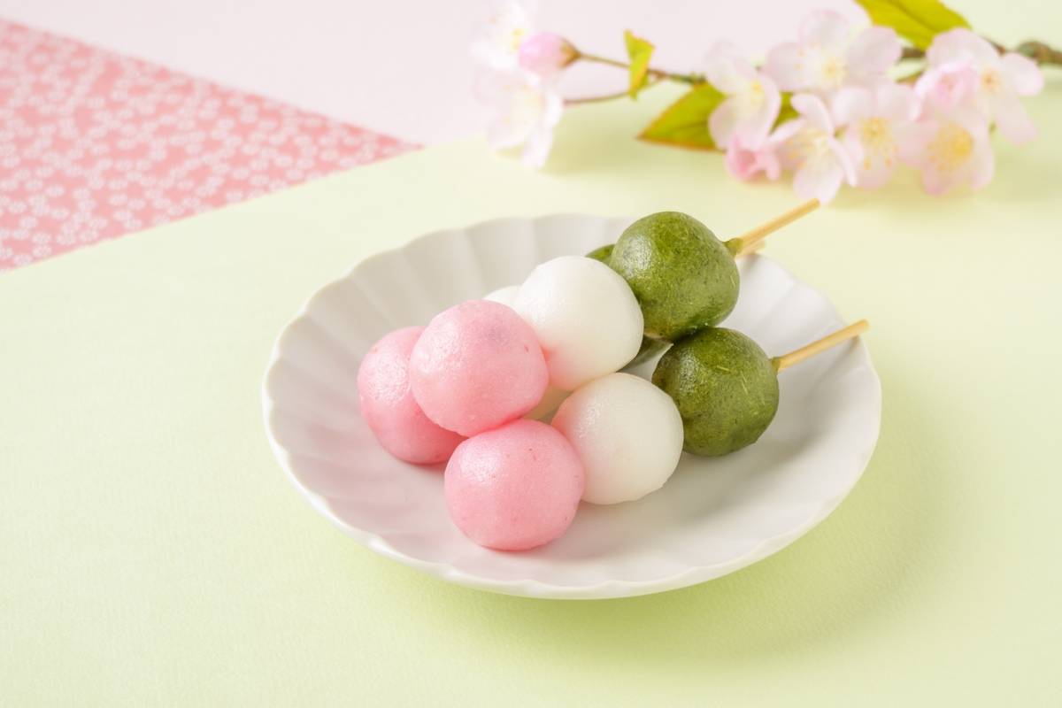 Three skewers of pink, white, and green dango on a white plate with cherry blossoms in the background.