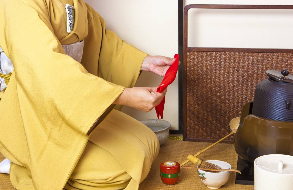 A person in a yellow kimono kneels, performing a tea ceremony with a red cloth and traditional tools.