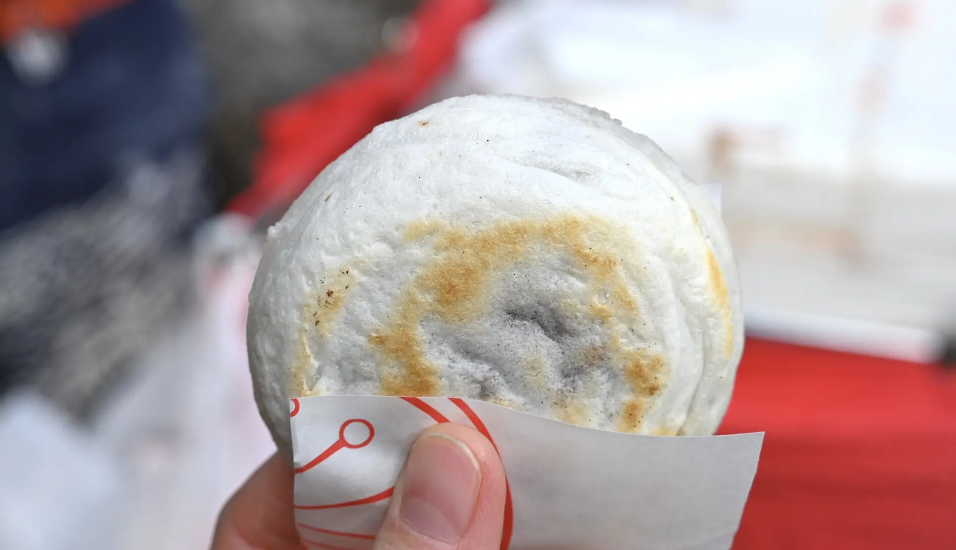 A hand holds a round, pan-fried bun with light brown spots, wrapped in white paper.