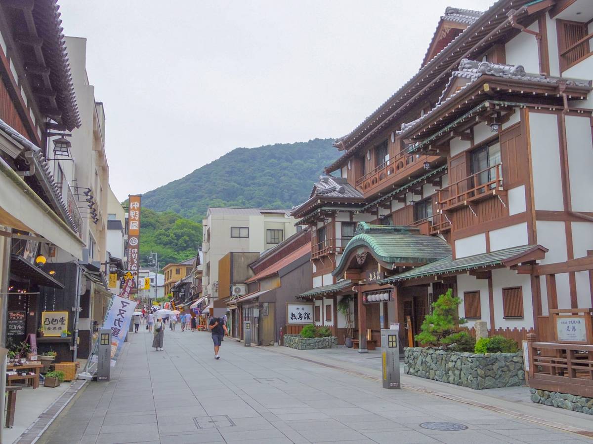 A street lined with traditional Japanese buildings, with people walking and a forested mountain in the background.